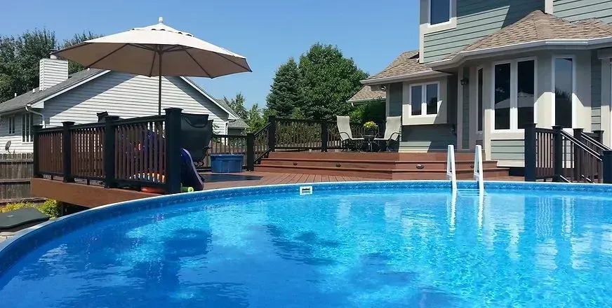 A large swimming pool with a deck and umbrella in front of a house.