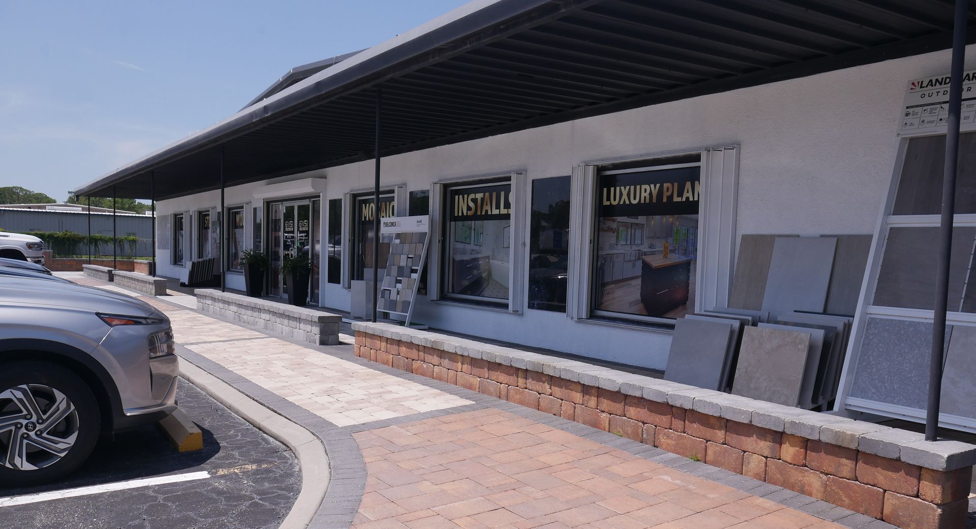 Exterior of a white building with black awning, storefront windows, and paving stones on display. A car is parked in front.