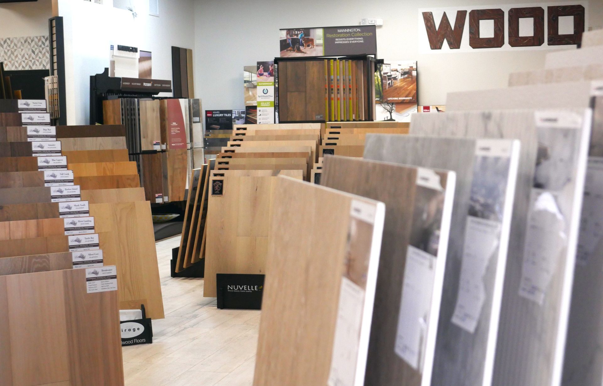 A showroom displays various wood flooring samples in different colors and styles, labeled with price tags. The word 