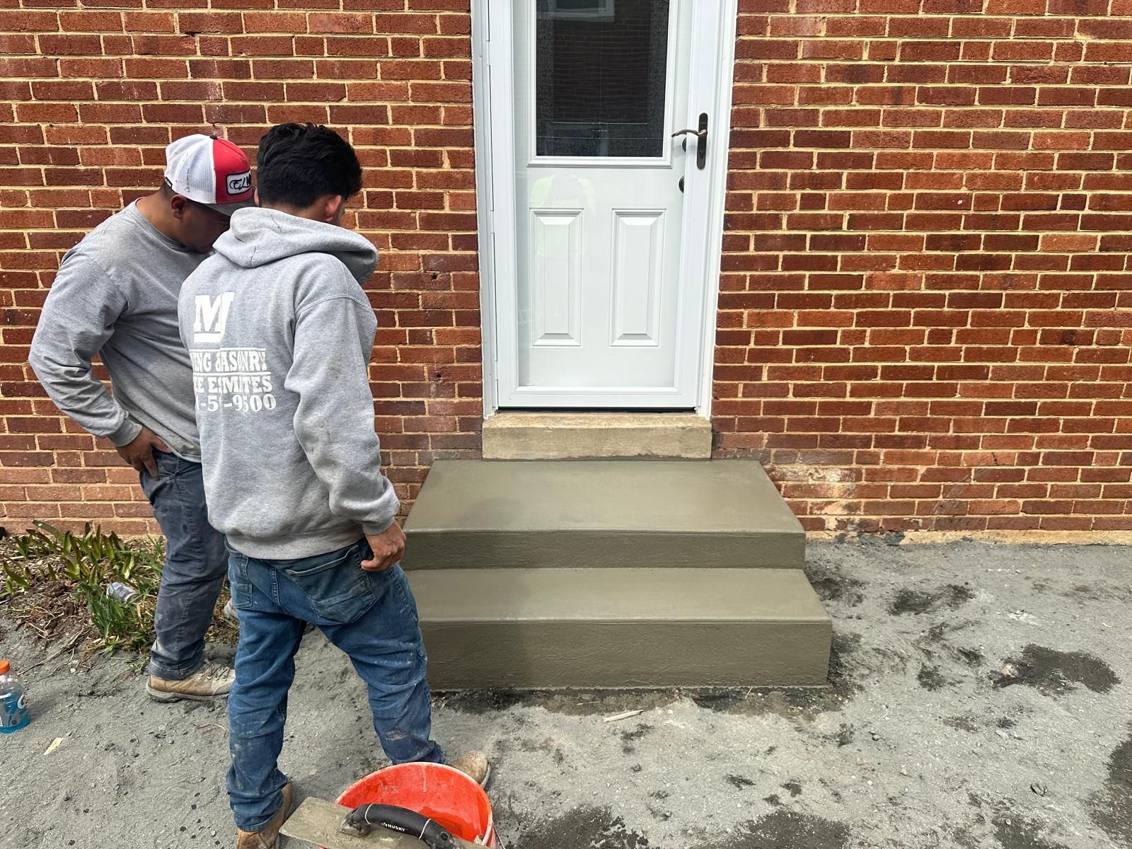 Two men are working on concrete steps in front of a brick building.