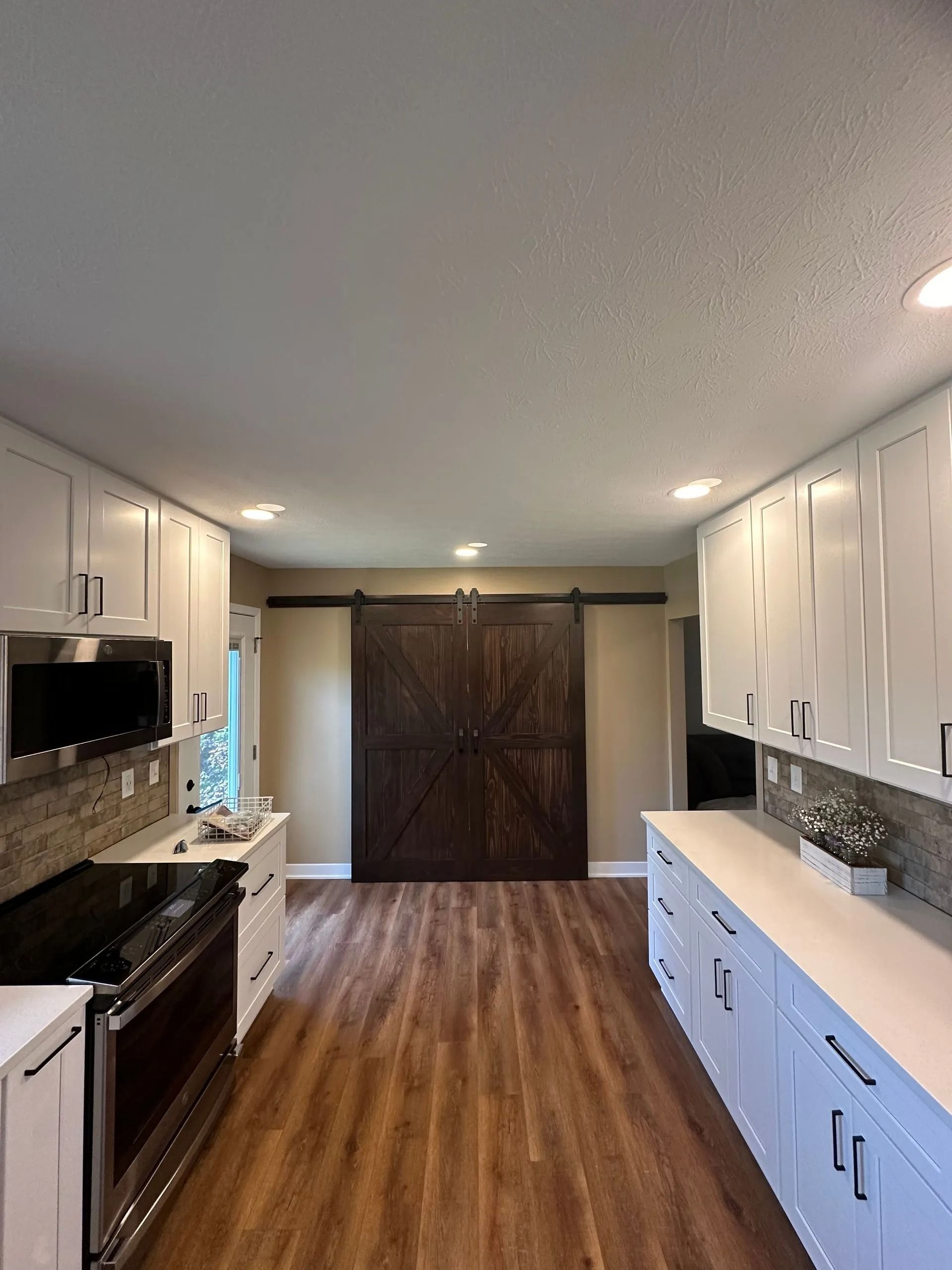 Modern kitchen with white cabinets, dark wood floor, and a large wooden double-door at the end.