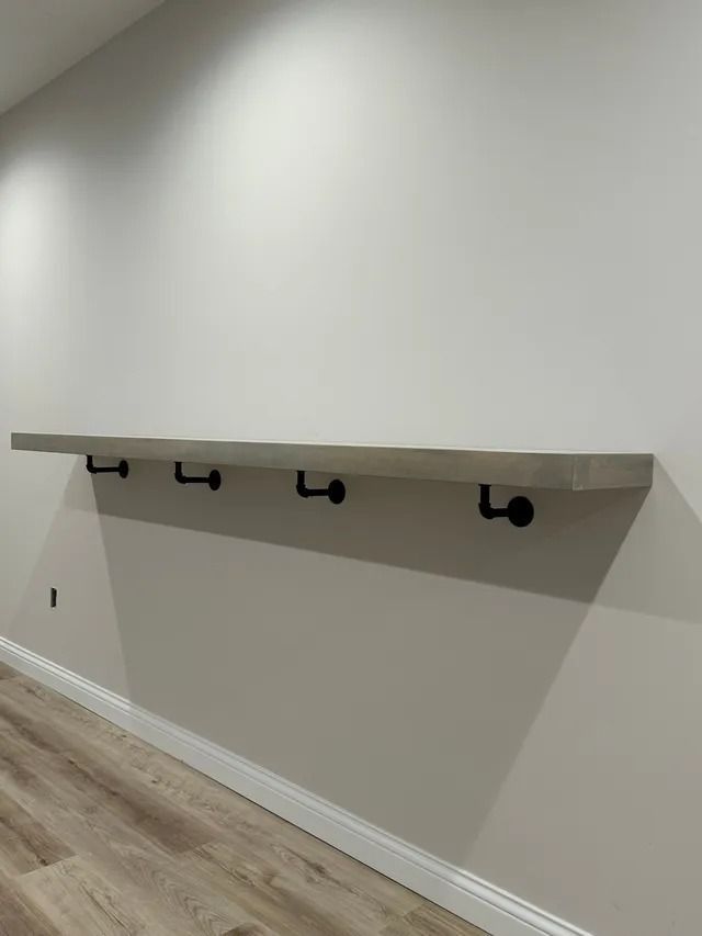 Empty hallway wall with a long wooden shelf and four black hooks above wood flooring