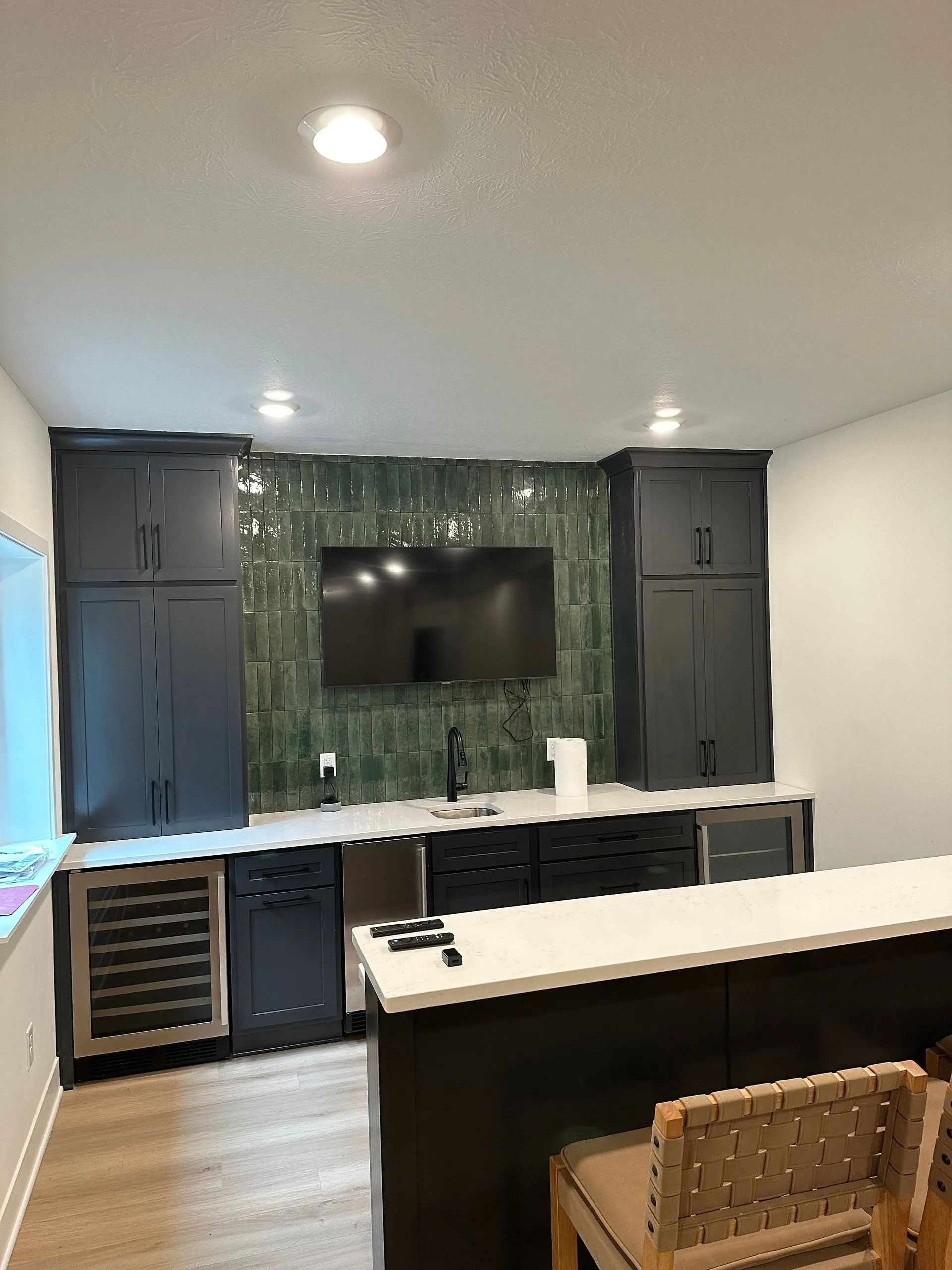 Modern kitchen with dark cabinets, green tile backsplash, wall-mounted TV, and white island countertop