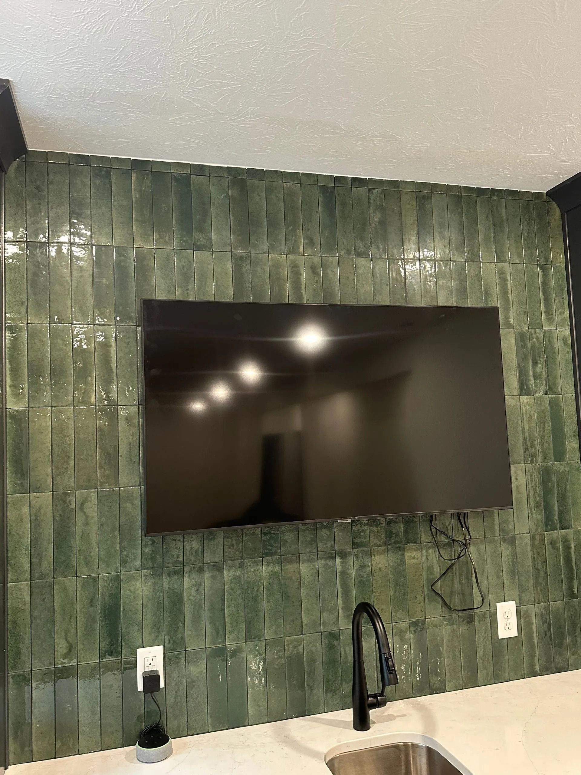Green tiled kitchen backsplash with a wall-mounted TV above a black faucet and white countertop.