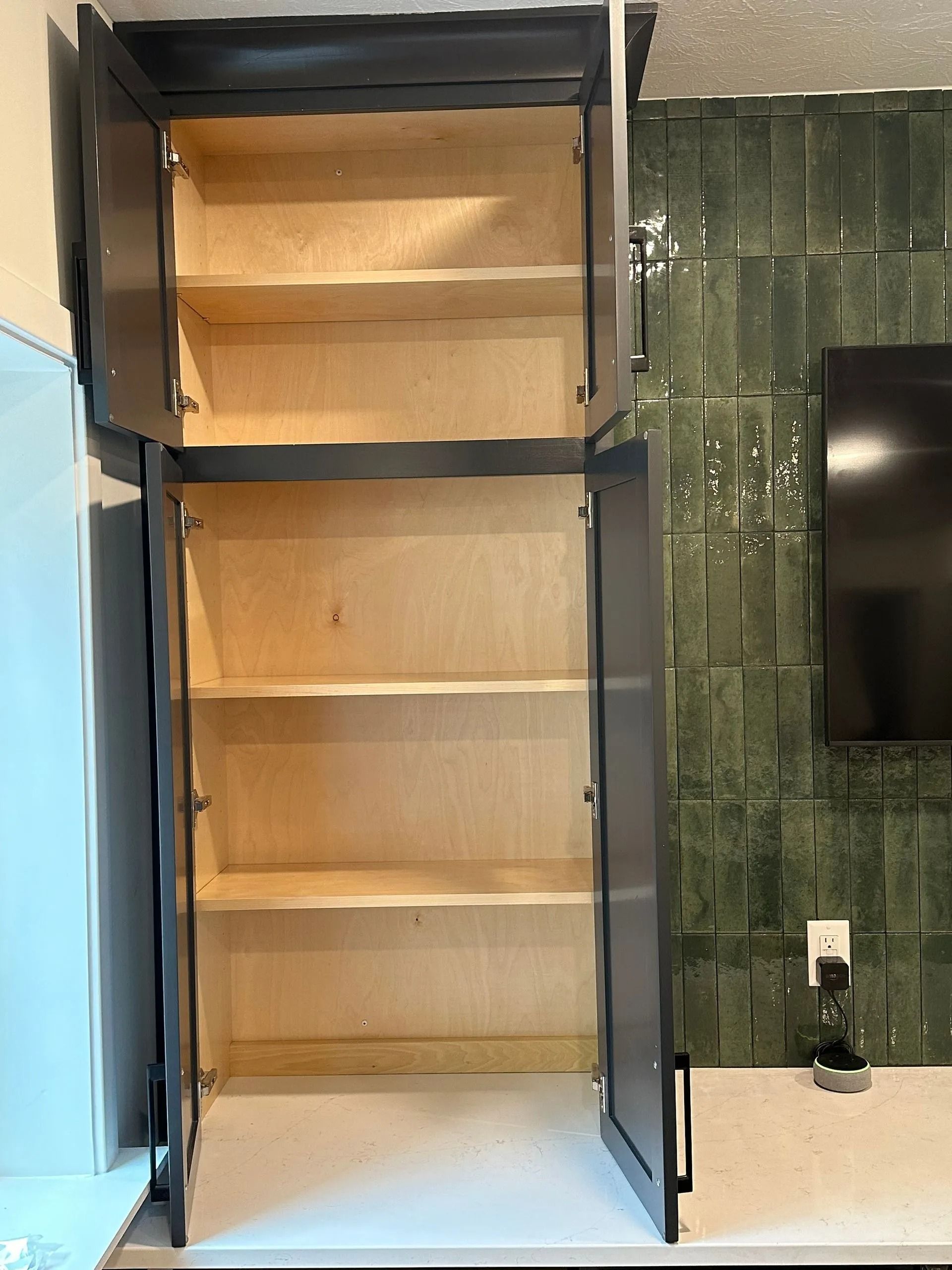 Open pantry cabinet with empty wooden shelves and dark frame beside green tiled wall.