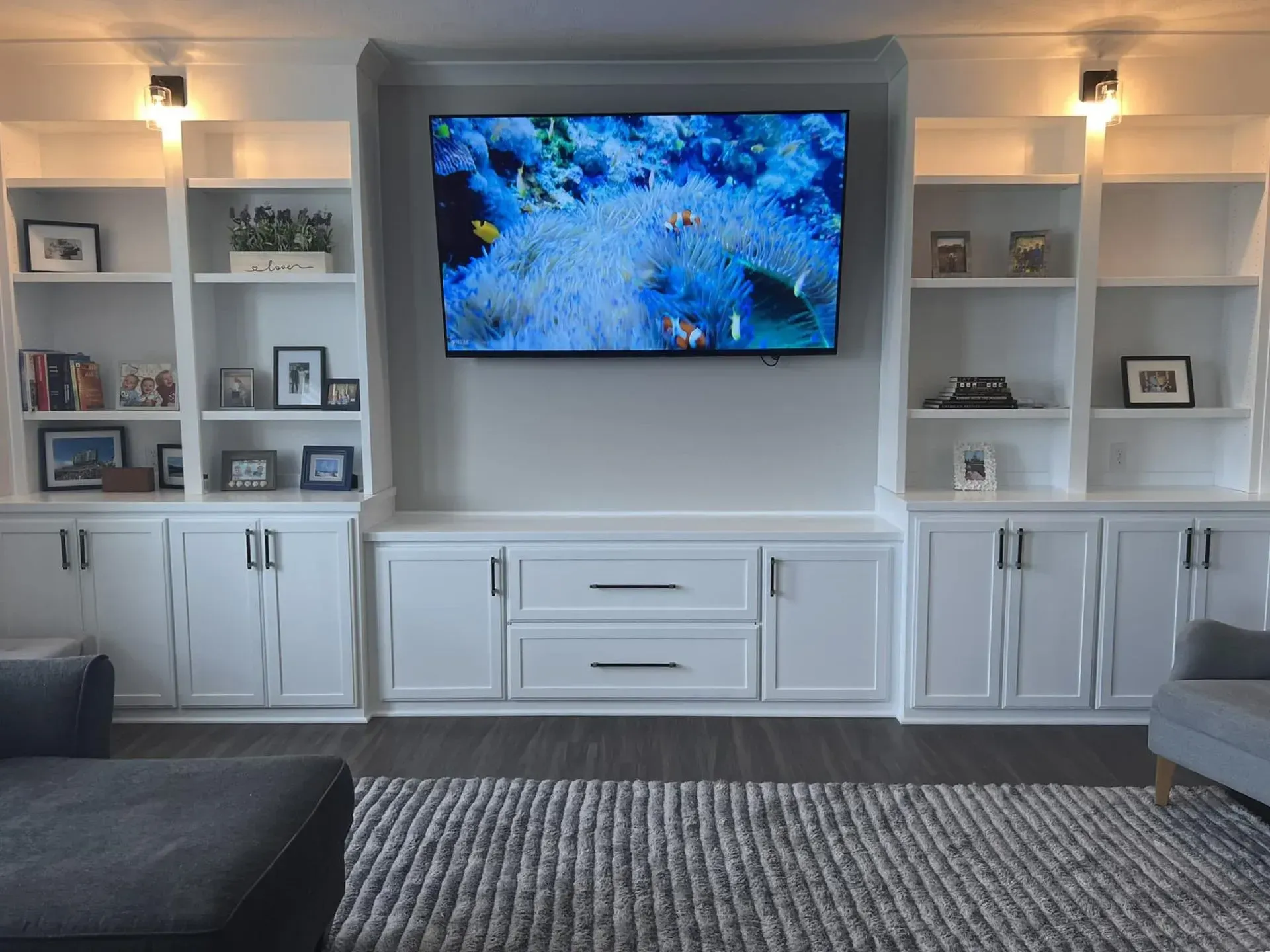 Living room wall with mounted TV, built-in shelves, cabinets, and gray ottomans on a patterned rug