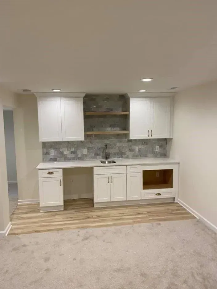 Empty basement kitchenette with white cabinets, gray tile backsplash, wood shelving, and light carpet flooring