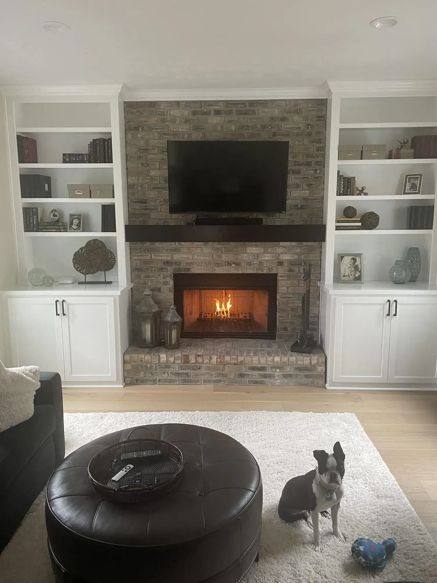 Living room with stone fireplace, built-in shelves, ottomans, and a small dog on the rug.