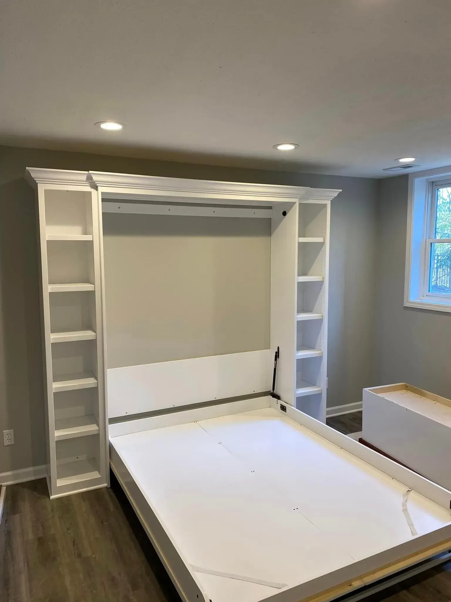 Wall-mounted white Murphy bed in a gray bedroom, with built-in shelves and a large cabinet frame.