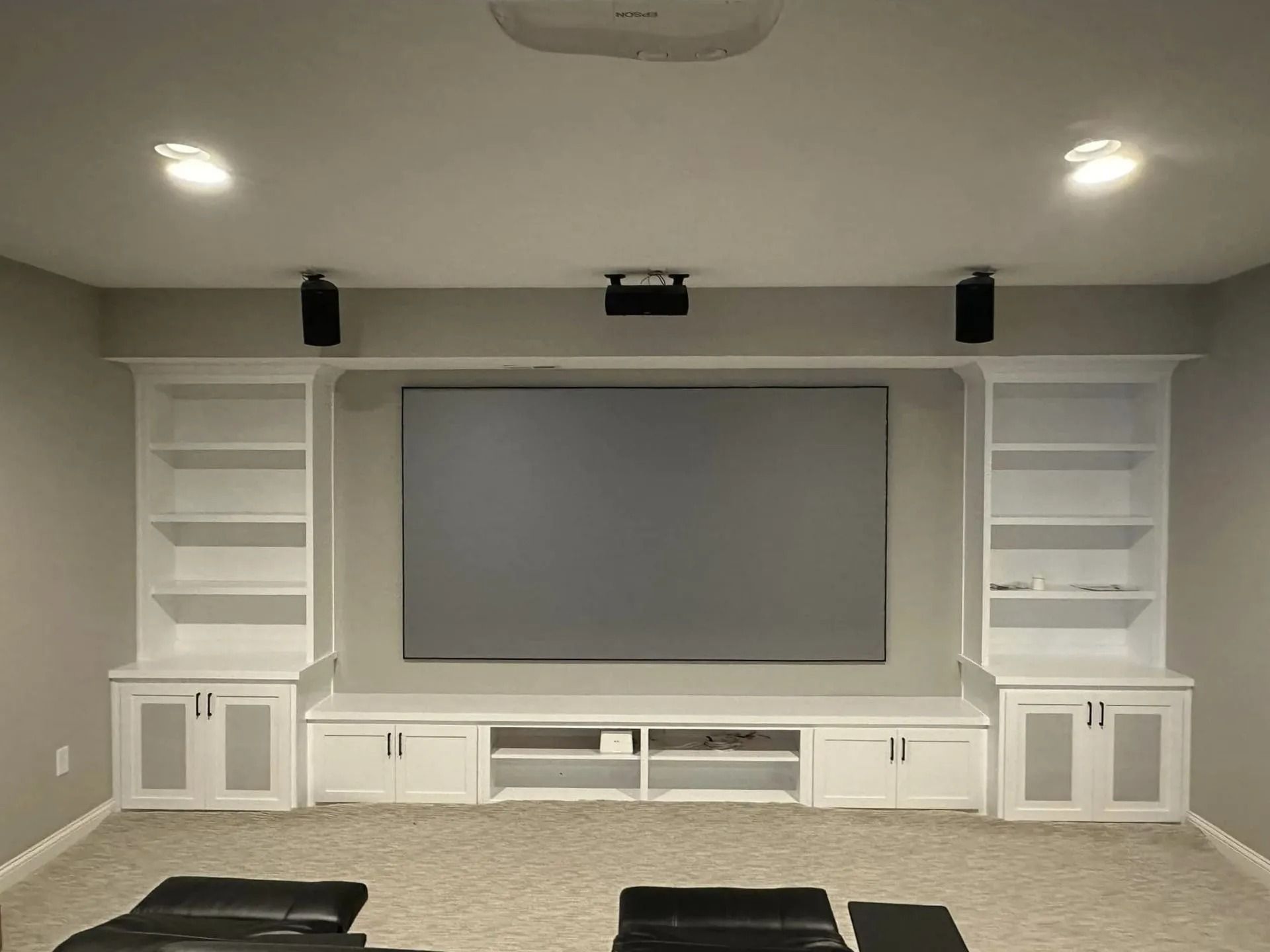 Home theater wall with built-in white shelves, cabinets, and a large gray projection screen under recessed lights