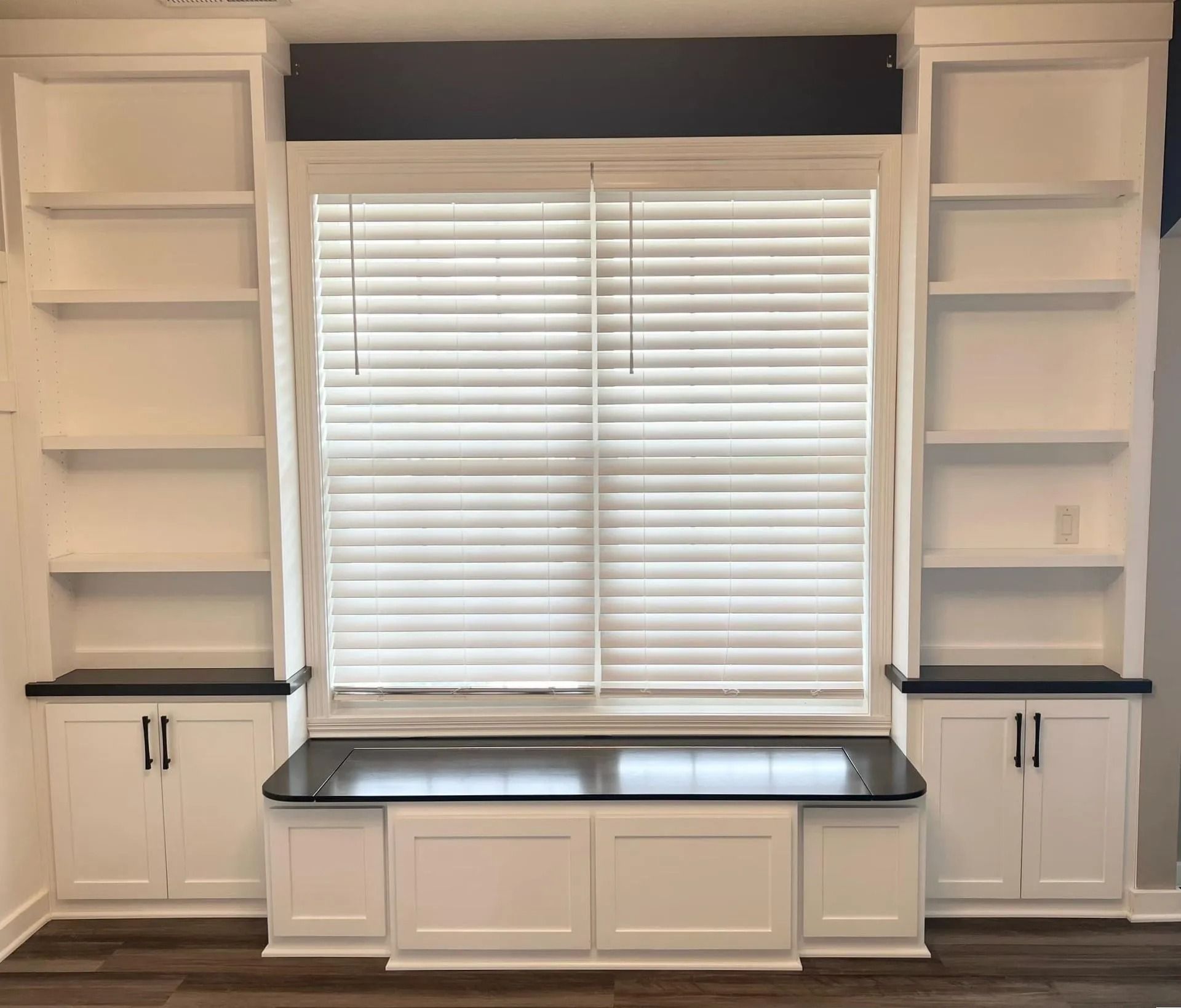 Built-in white shelving and cabinets framing a window with blinds and a dark countertop bench