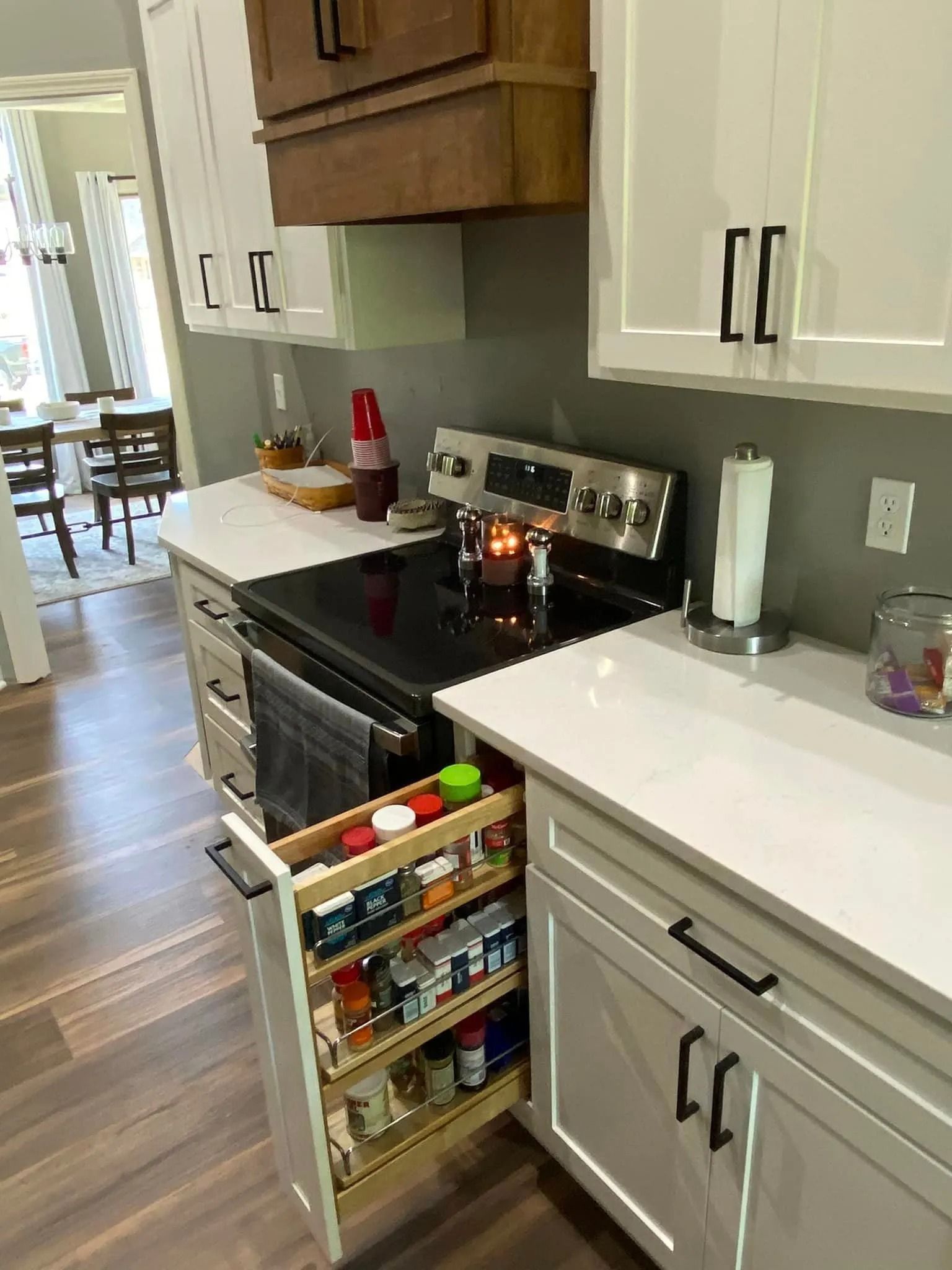 Kitchen with open oven drawer, stovetop, white cabinets, and a pull-out pantry stocked with jars and cans.