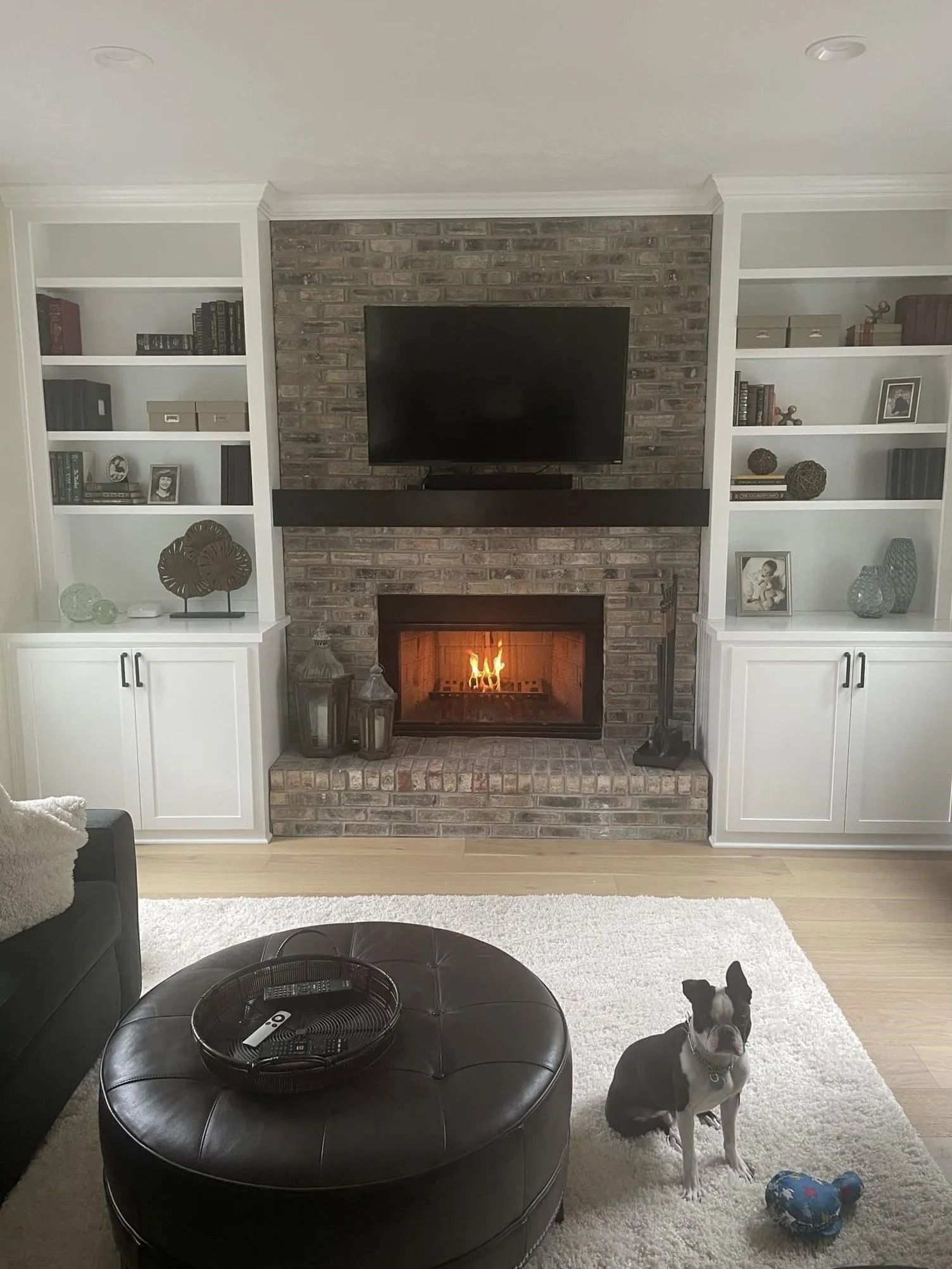 Cozy living room with brick fireplace and TV, white built-ins, round ottoman, and a small dog on the rug