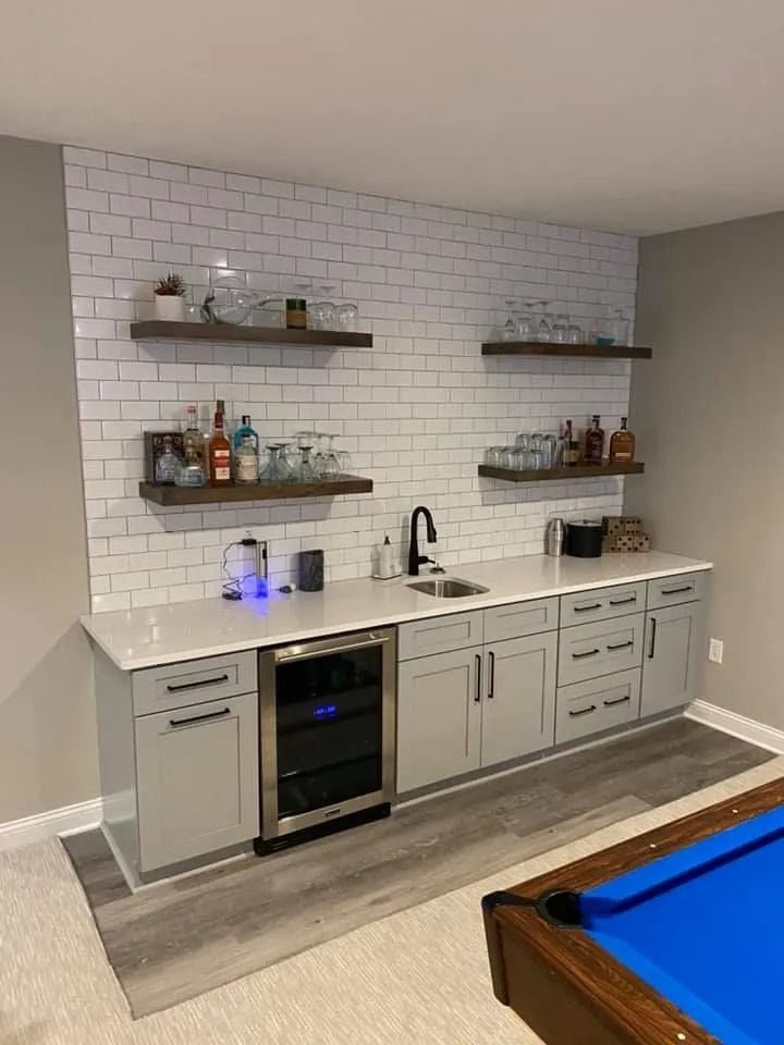 Modern basement wet bar with white tile backsplash, floating shelves, and blue pool table in foreground
