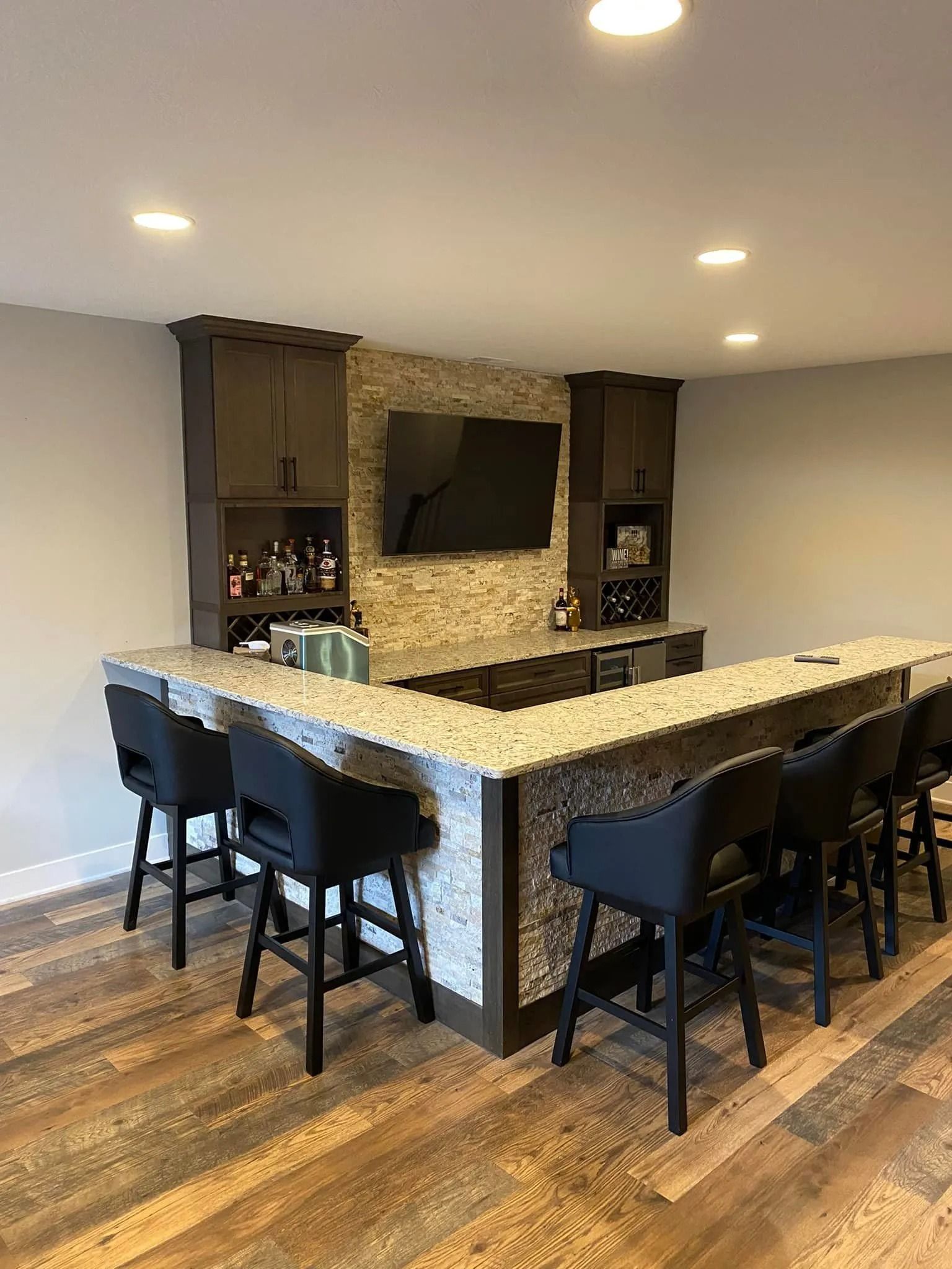 Modern home bar with granite countertop, black stools, stone accent wall, and wall-mounted TV