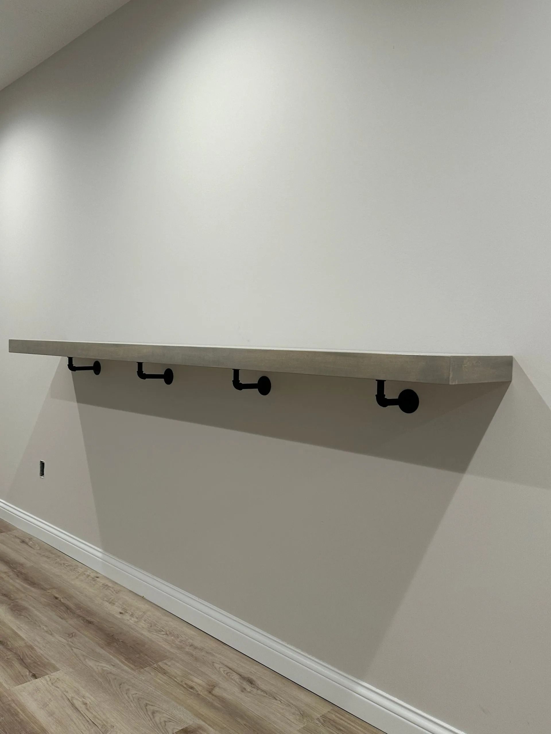 Wall-mounted wooden shelf with black metal brackets in an empty room with light walls and wood flooring