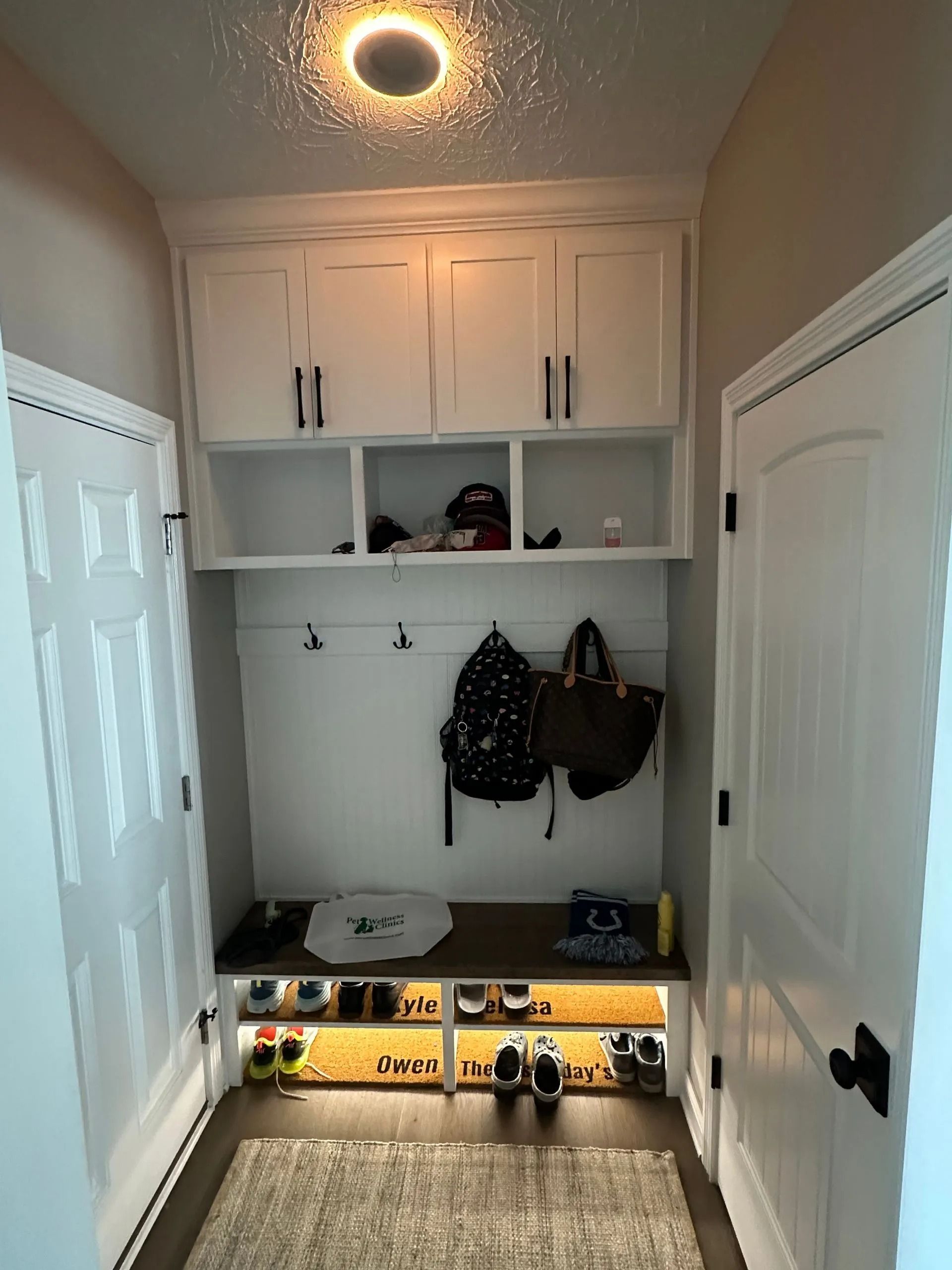 Small entryway mudroom with white cabinets, hooks, bags, and shoe storage with warm under-shelf lighting.