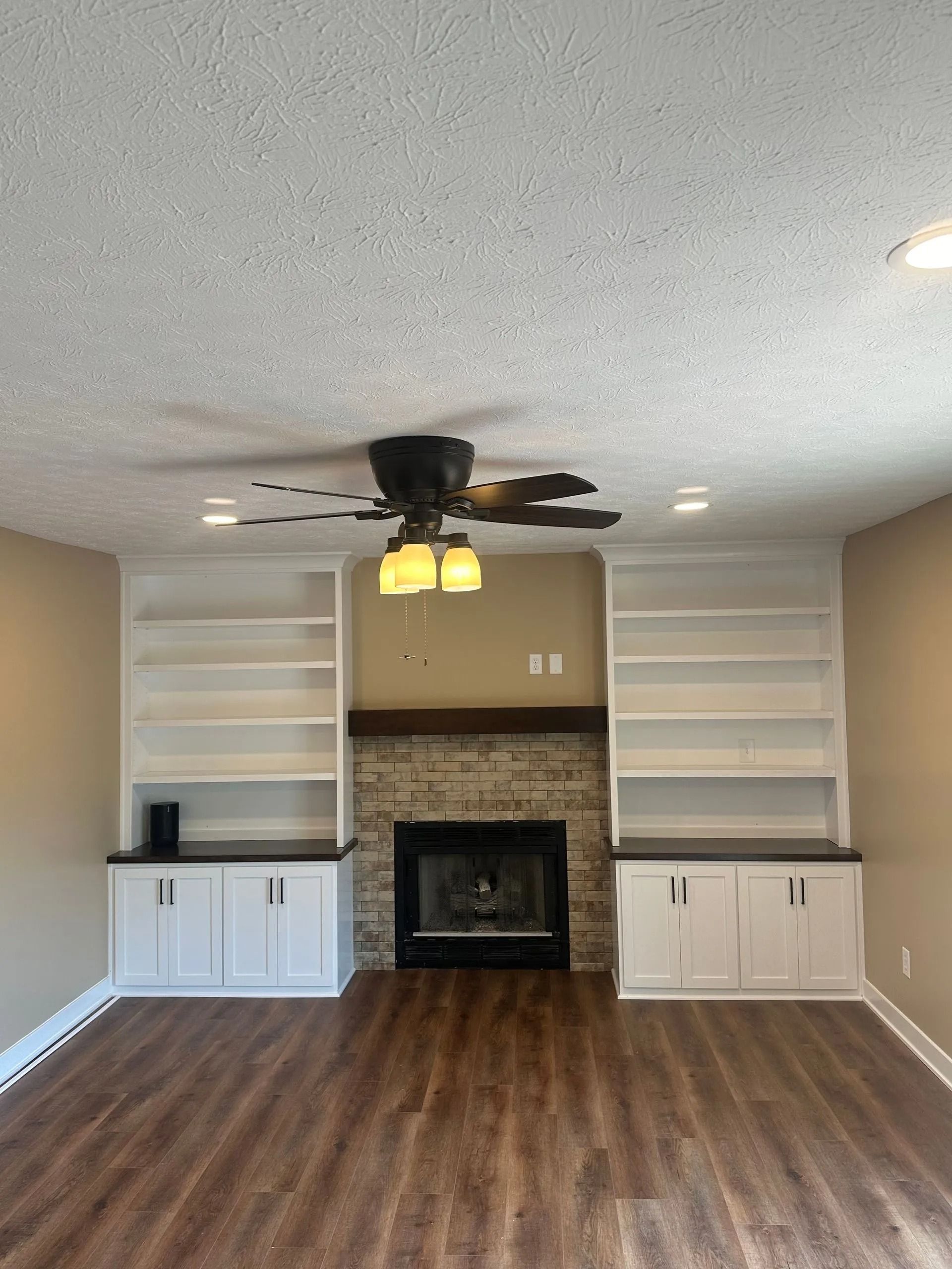 Empty living room with built-in shelves, fireplace, ceiling fan, and wood-look flooring.