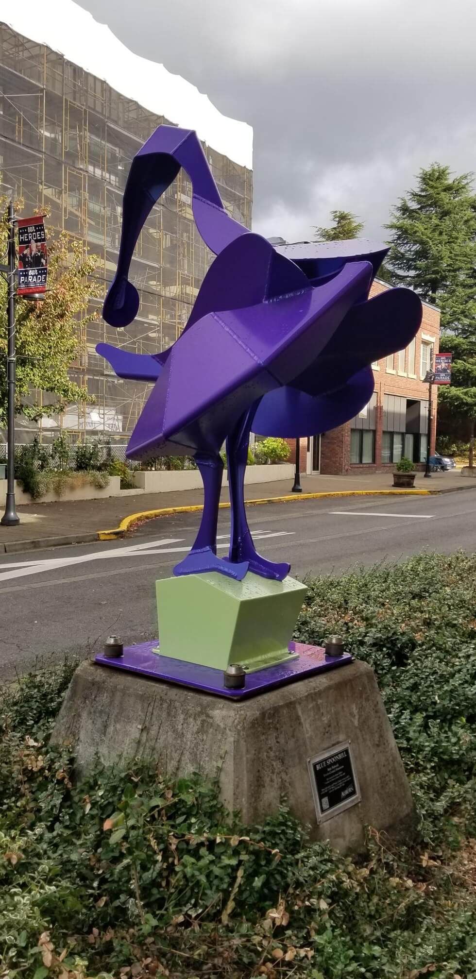 Purple metal sculpture of a bird-like figure on a concrete base, located in an outdoor urban setting.