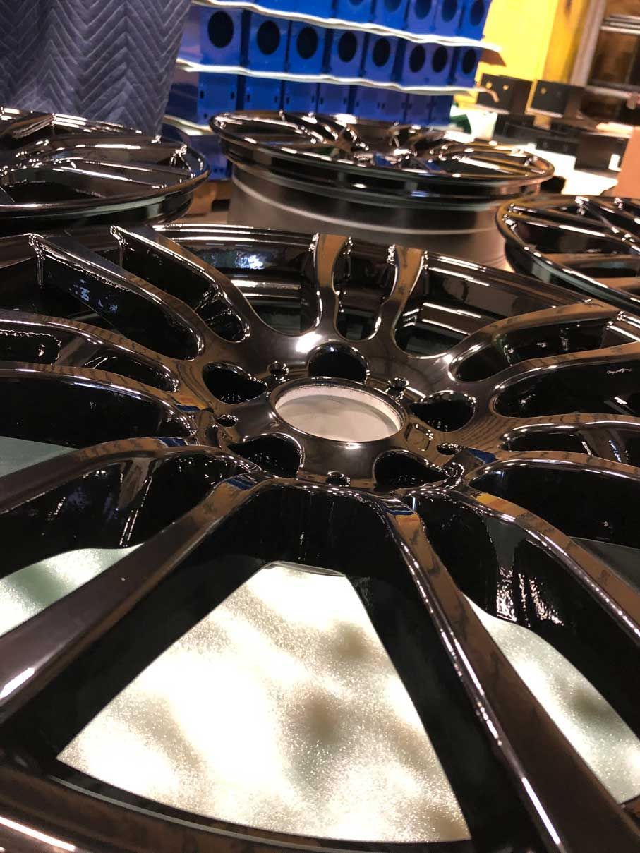 Black alloy car wheels, multiple, in a workshop. Shiny finish, with hubcaps and other parts in the background.