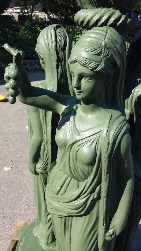 Green statue of a woman holding grapes, wearing draped clothing. Another figure is in the background.