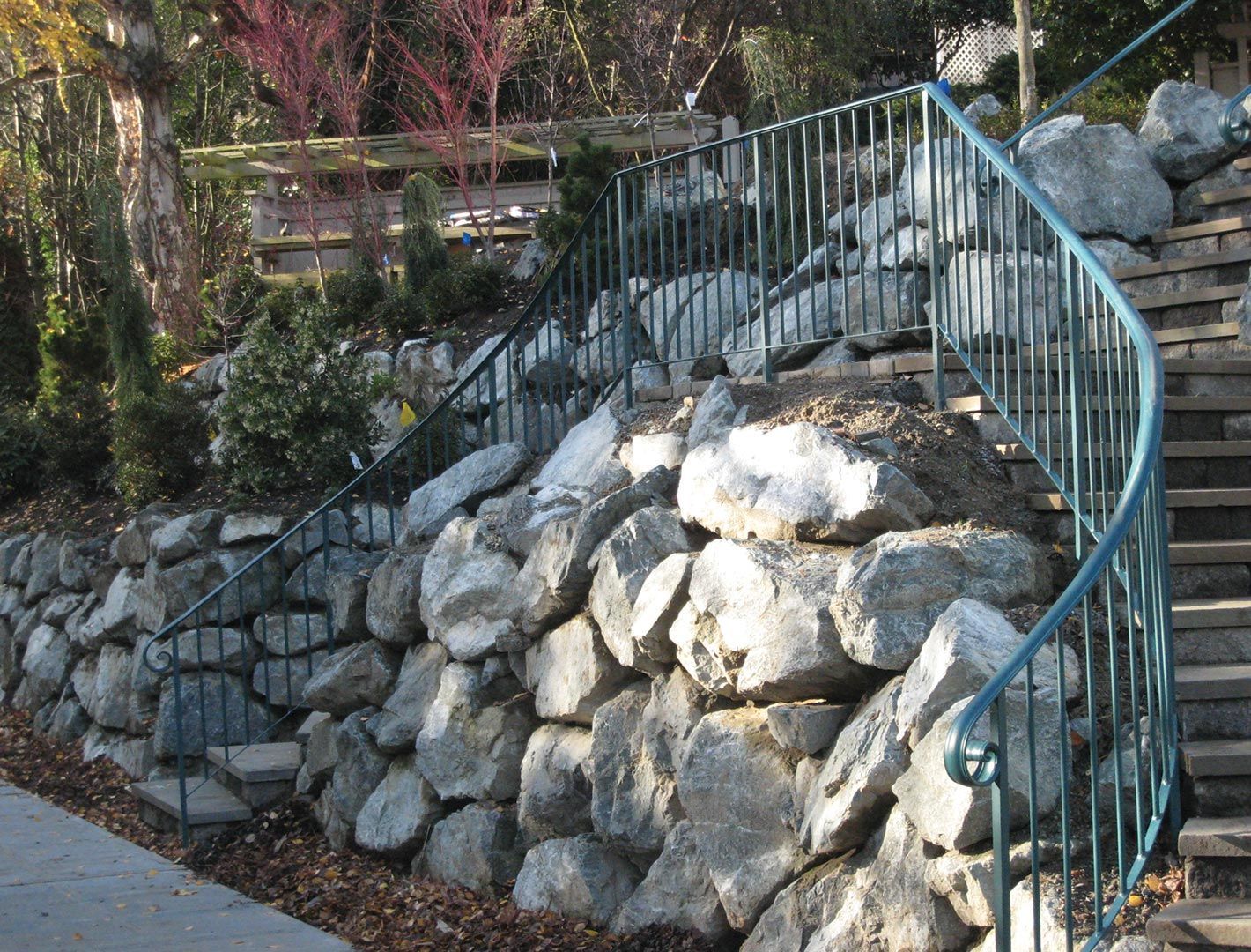 Stone retaining wall with a curved blue metal handrail and steps leading upward.