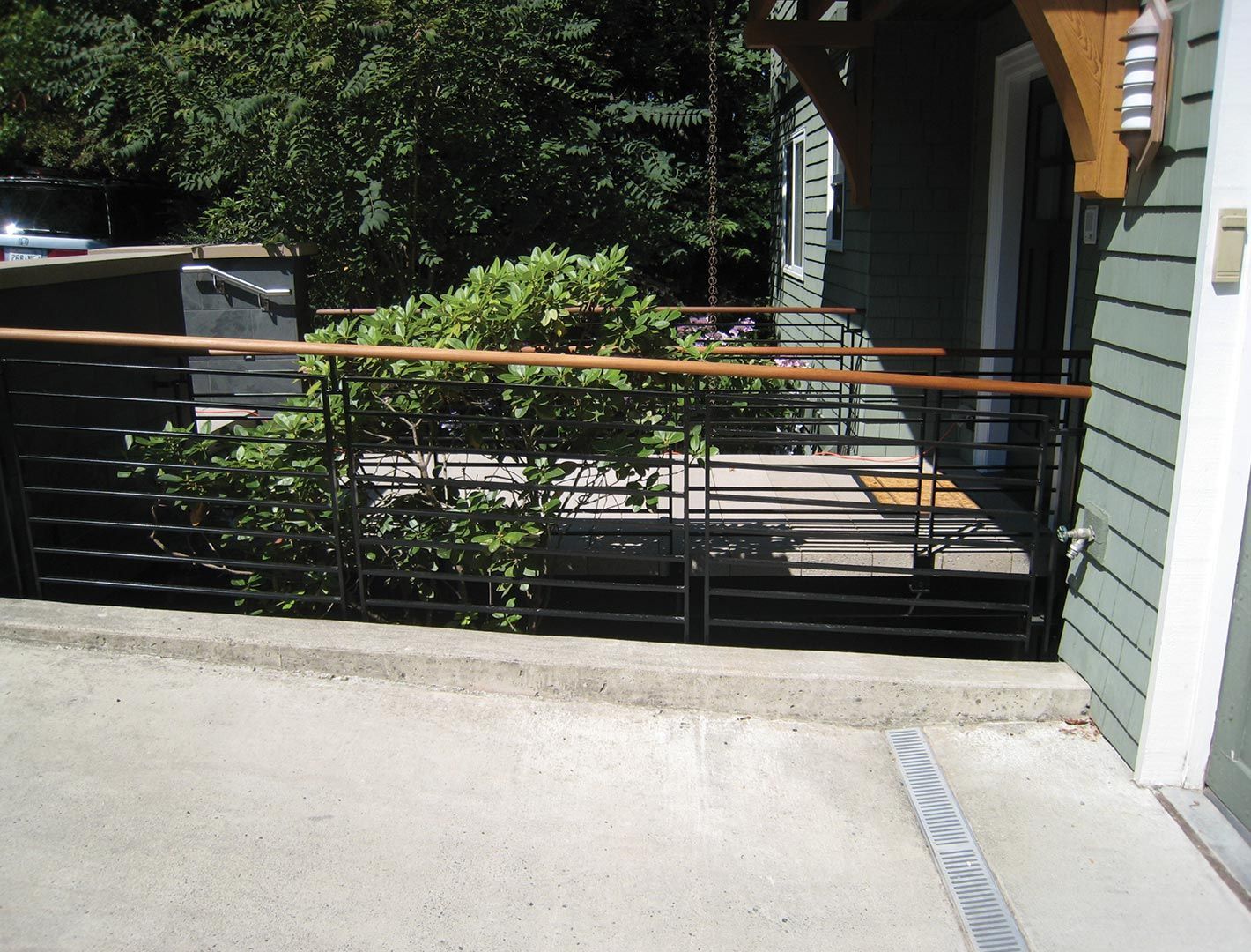 A low concrete patio with black metal railing and wooden handrail, in front of a green building and lush greenery.