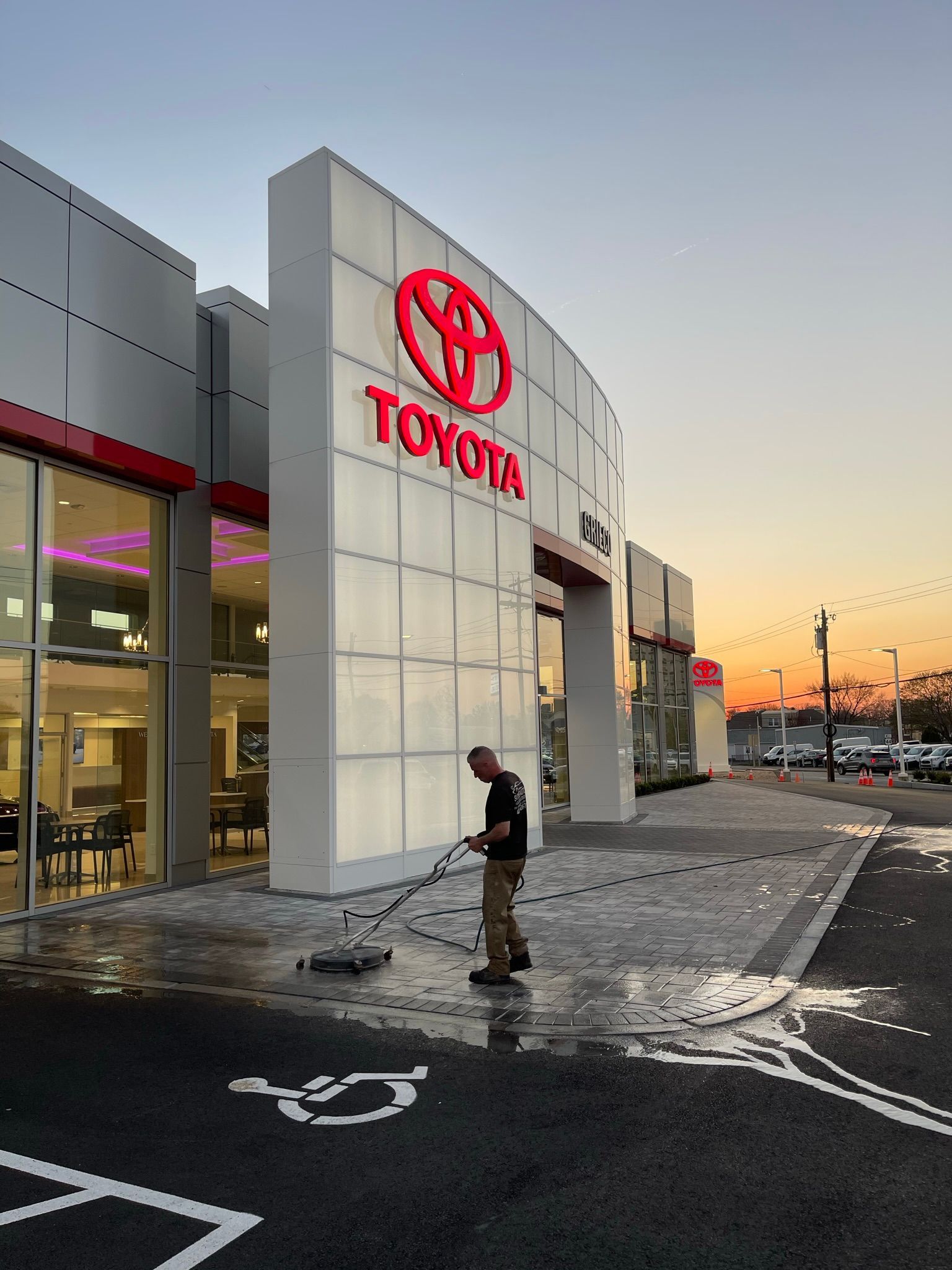 Man power washes the sidewalk in front of a Toyota dealership with a large red logo, at dusk.