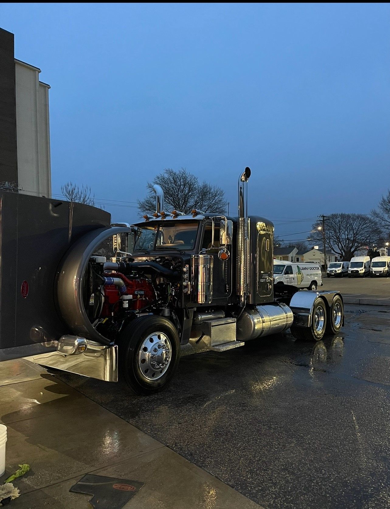 Semi-truck with open engine bay parked outdoors. Chrome accents, gray body, wet pavement, overcast sky.