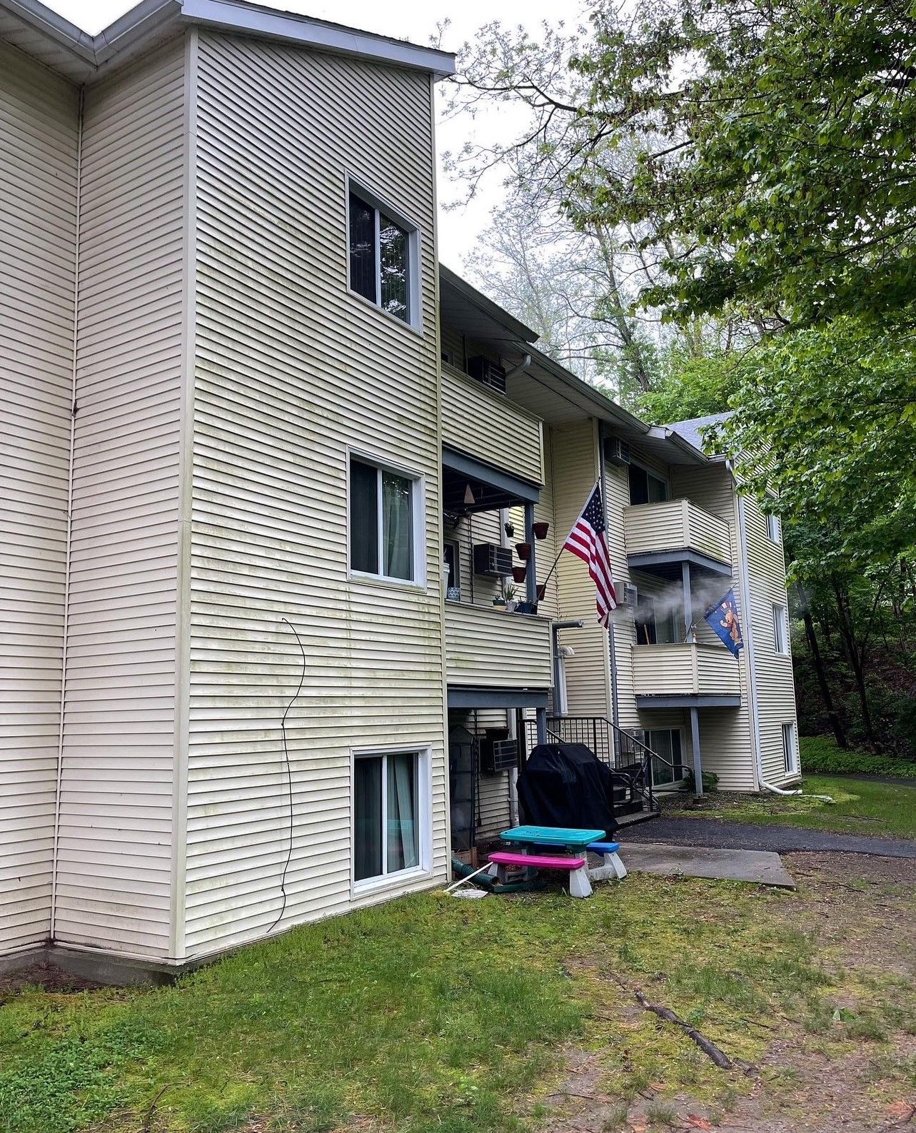 Two-story apartment building with light-colored siding, windows, and flags. Green grass and trees are in the foreground.