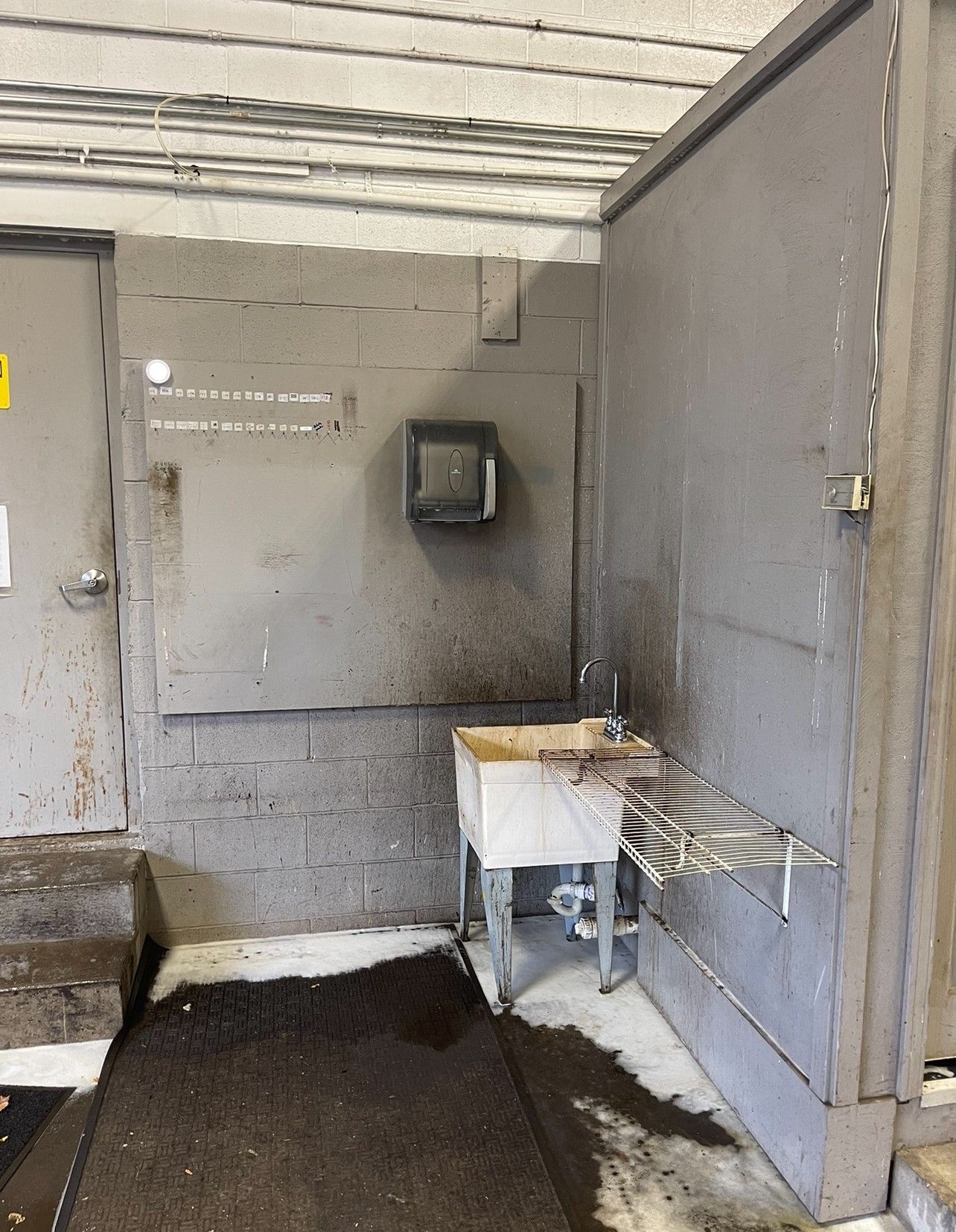 Dirty industrial wash station; sink, hand dryer, door. Dark, soiled, and potentially hazardous environment.