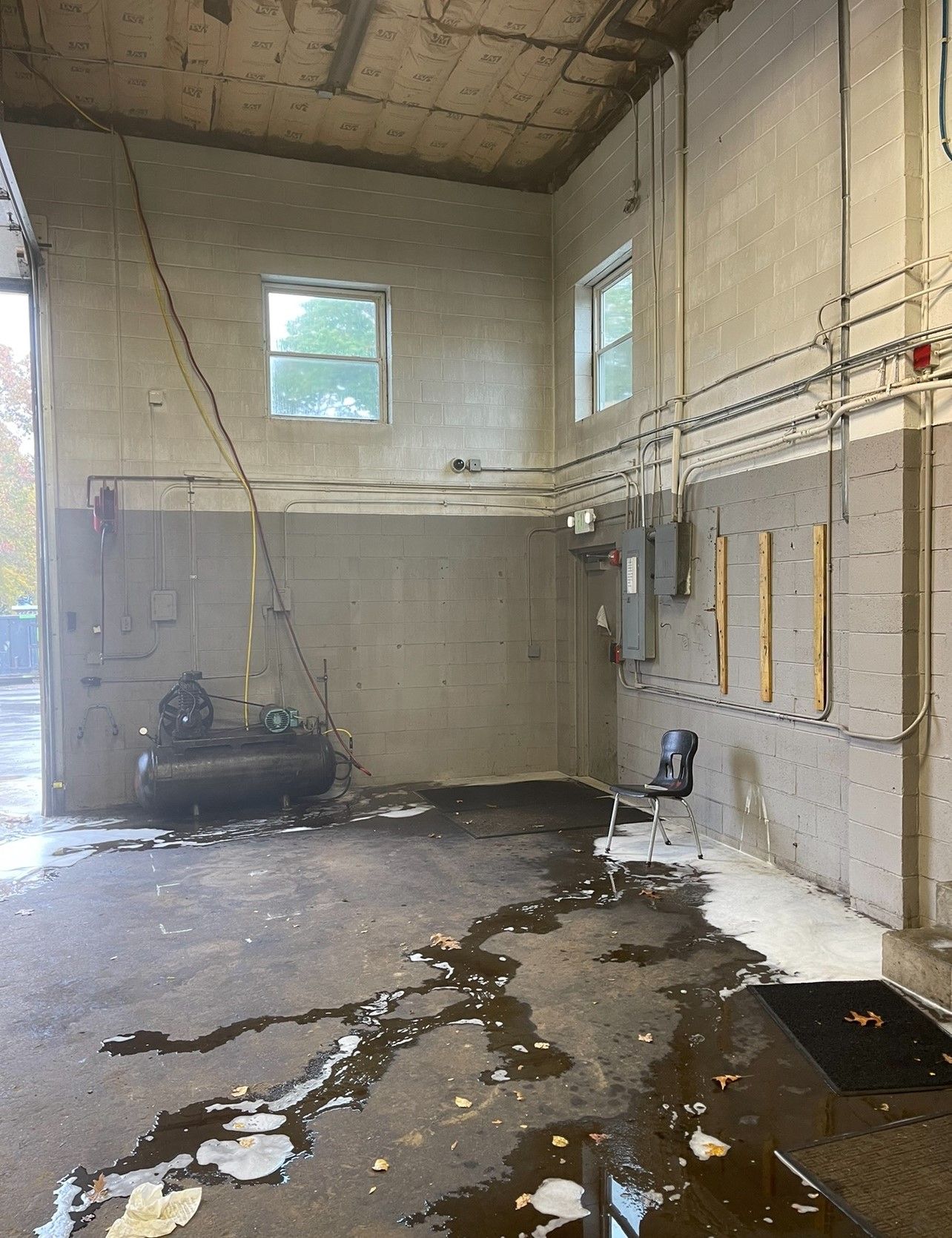 Interior of a garage with wet floor. Block walls, two windows, wiring, and a black chair are visible.