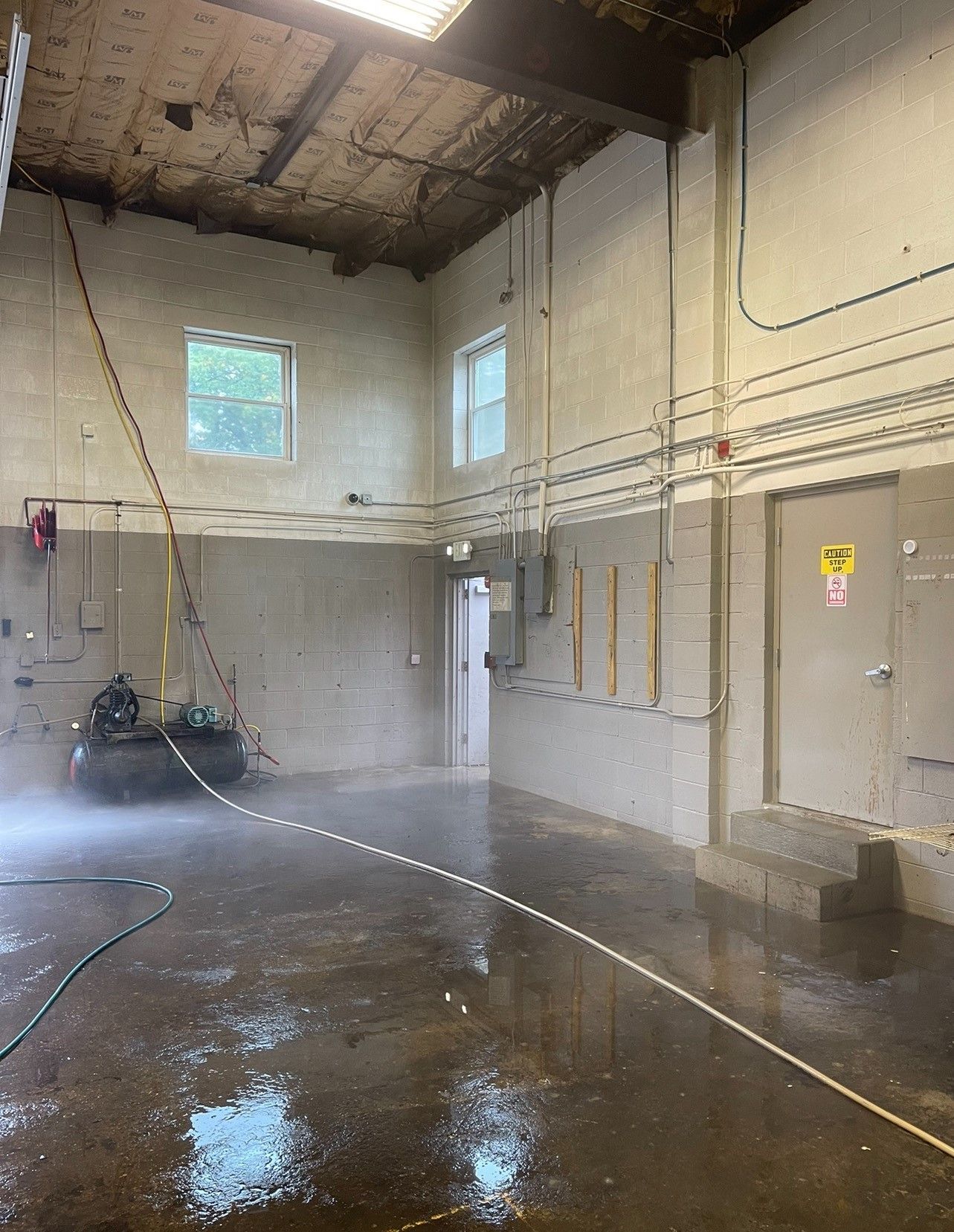 Interior view of a wet, empty room being sprayed with water. Concrete walls, two windows, and a doorway are visible.