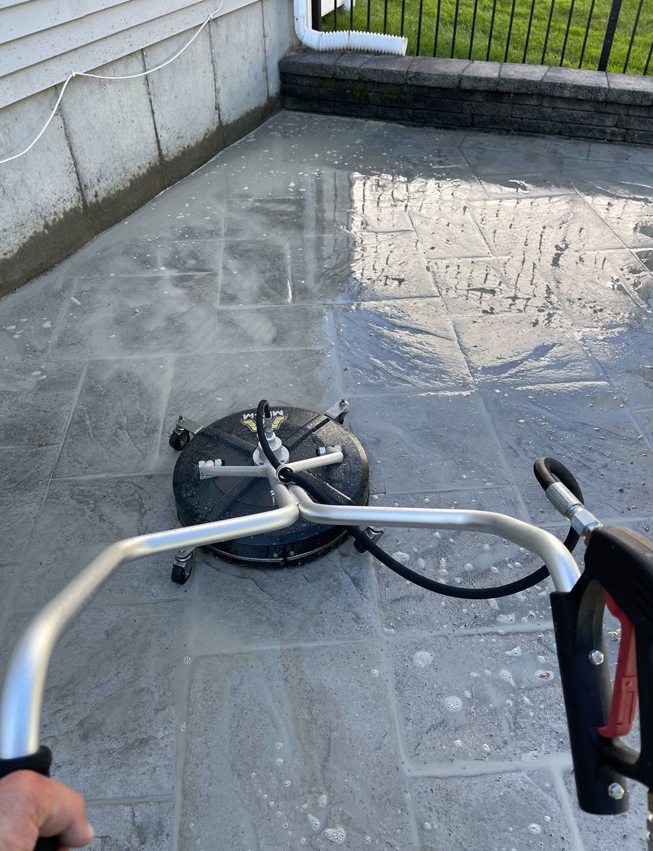 A person uses a surface cleaner to wash a gray stone patio near a building.
