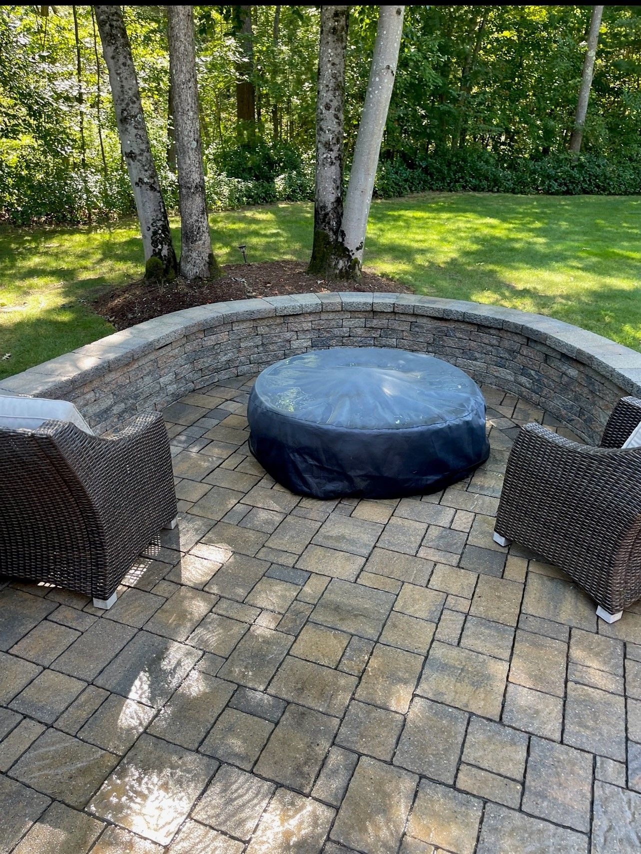 Patio with fire pit covered in black, surrounded by curved stone wall and two wicker chairs.