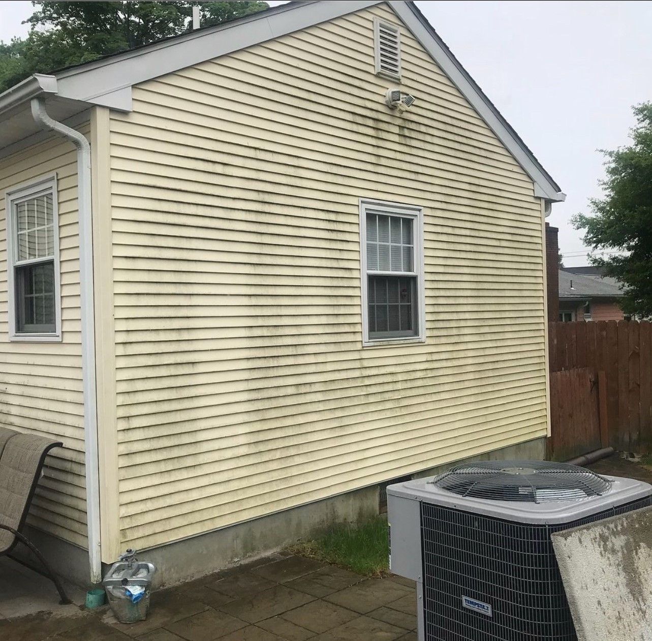 Yellow-sided house with two windows, a small air conditioner, and a bench on a patio.