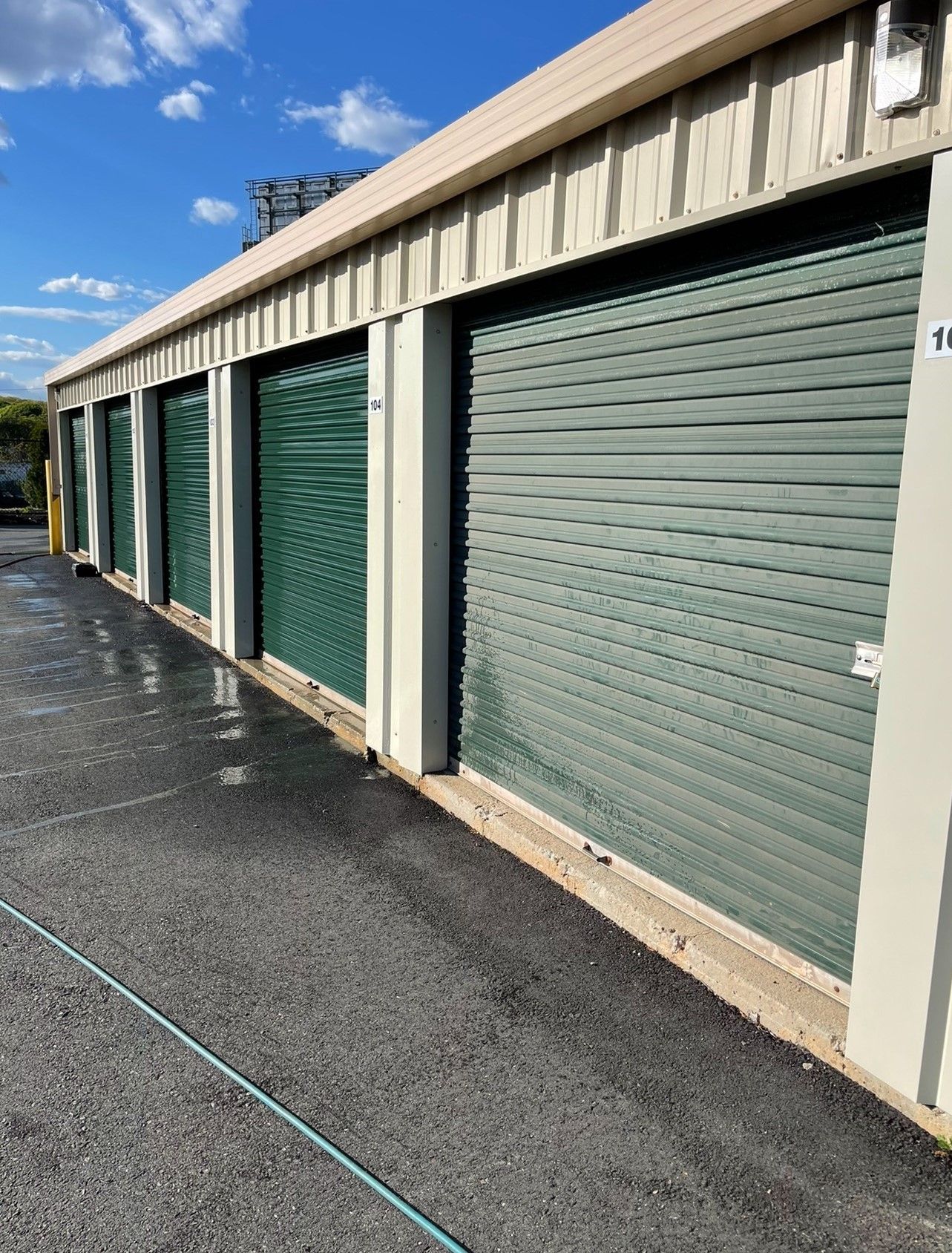 Storage units with green roll-up doors, along a black asphalt walkway, under a blue sky.