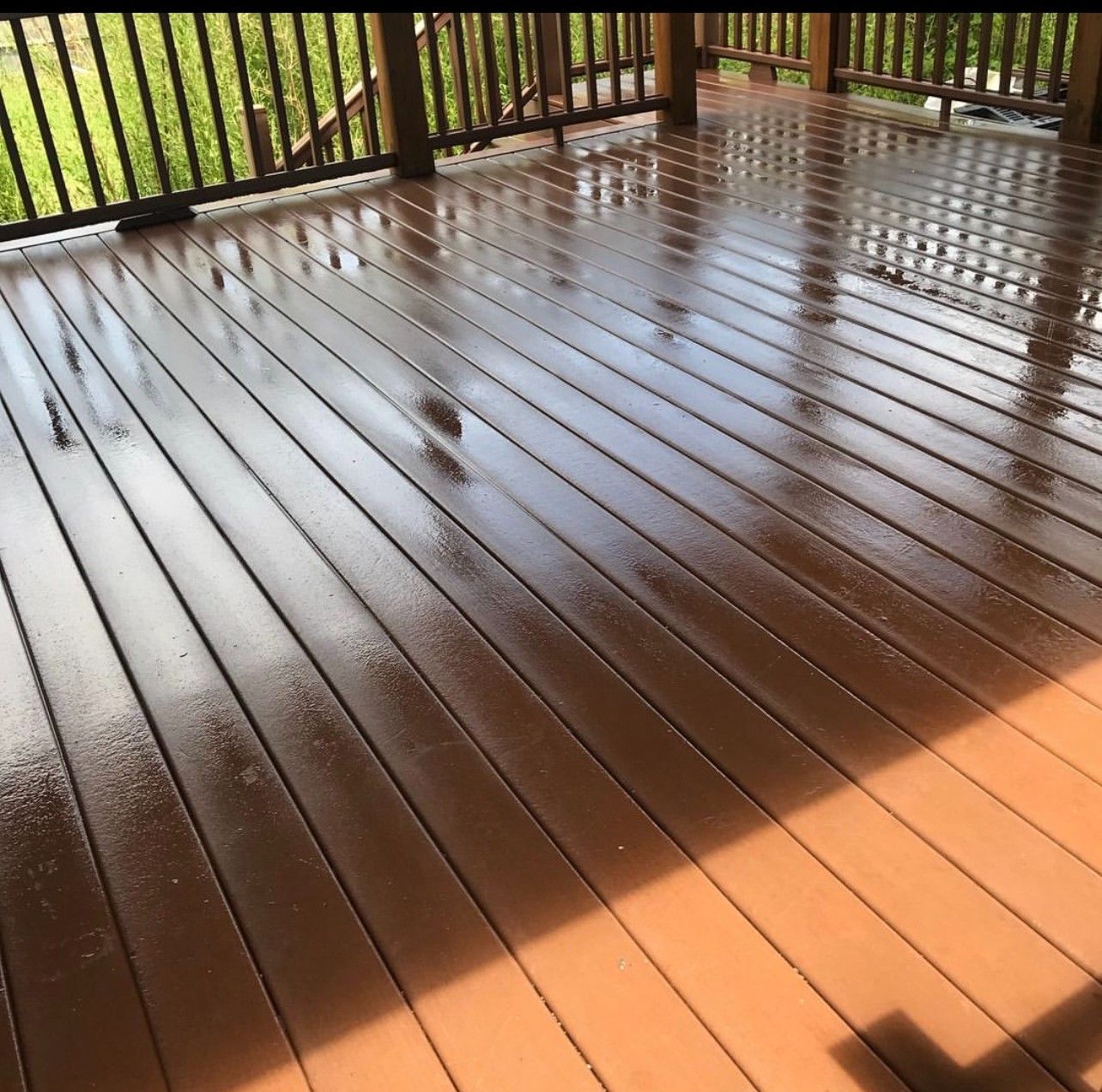 Wooden deck with a glossy brown finish, reflecting sunlight. Dark railing and green grass in the background.