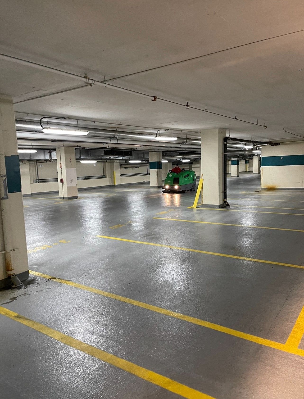 Green utility vehicle in a mostly empty parking garage with wet, gray floors and yellow parking lines.