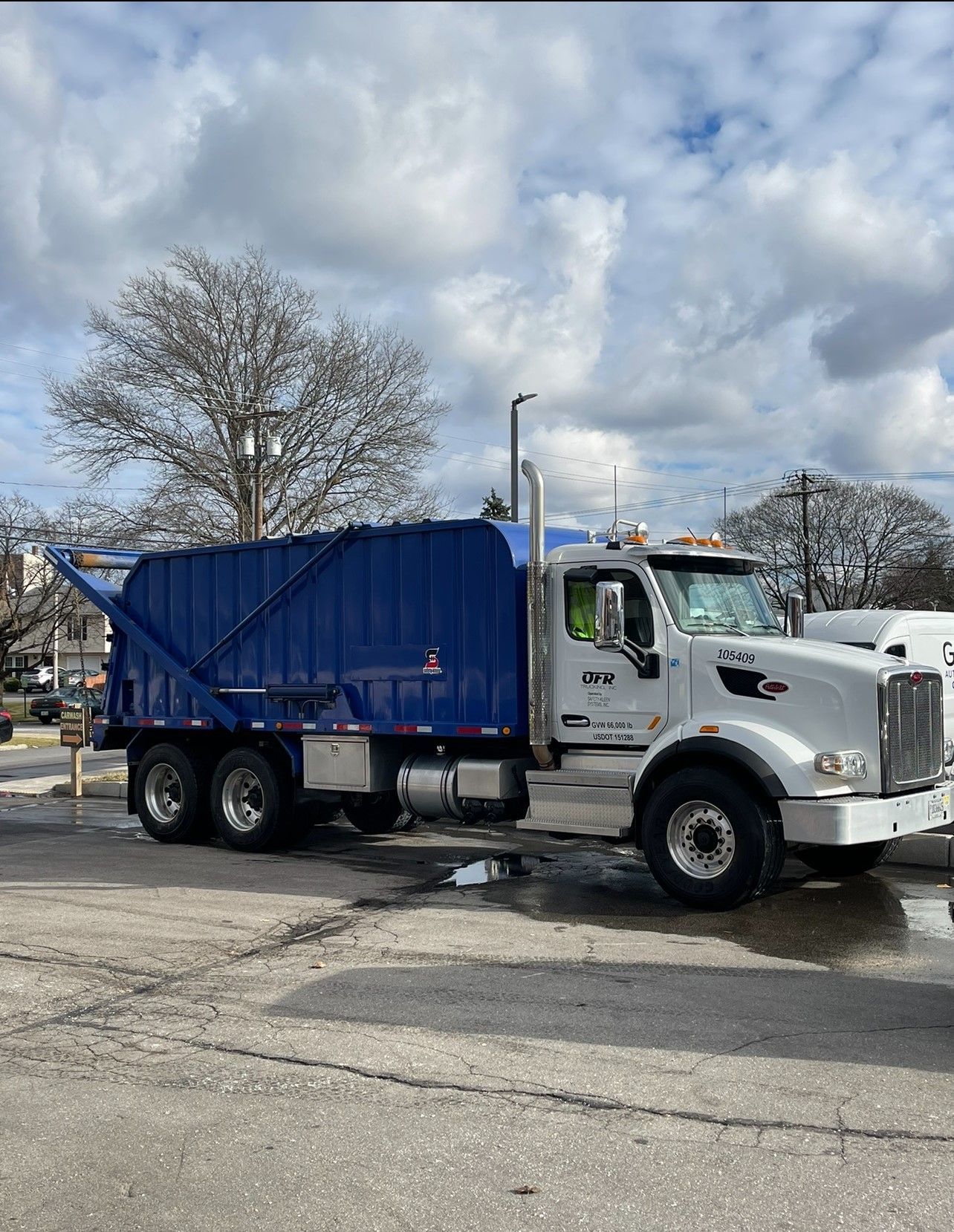 Blue and white garbage truck parked on a paved lot with a cloudy sky.