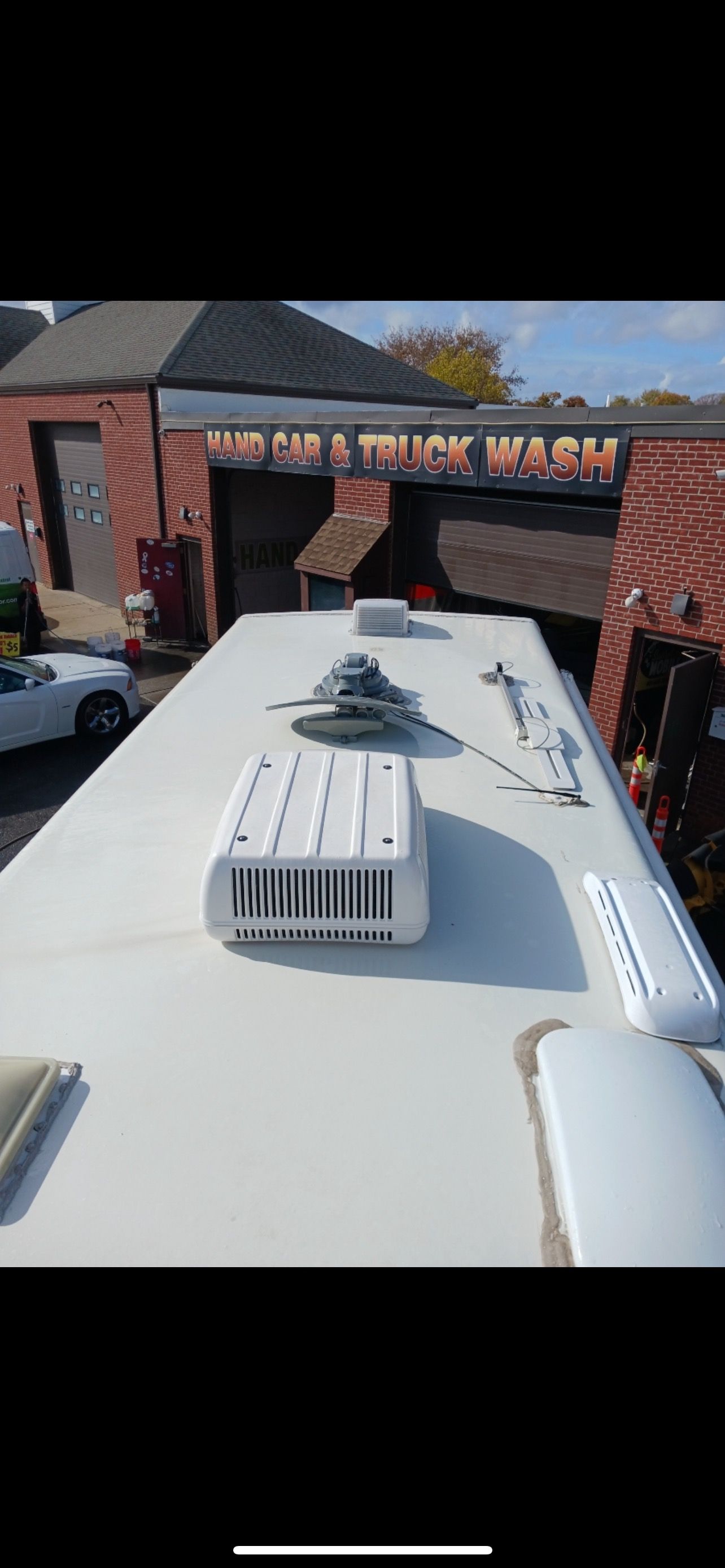 View from above, a white RV roof with vents and air conditioning unit. Brick buildings and trees in the background.