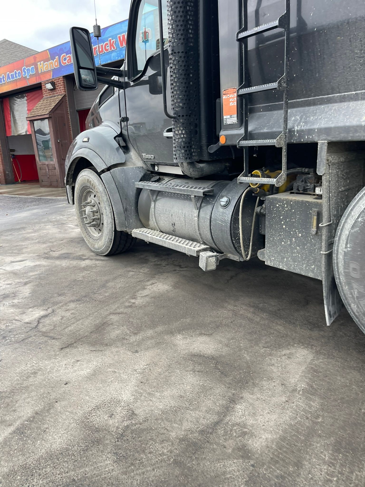 Black semi-truck parked on a gray, dirty surface. The truck has a fuel tank, step, and tire visible.