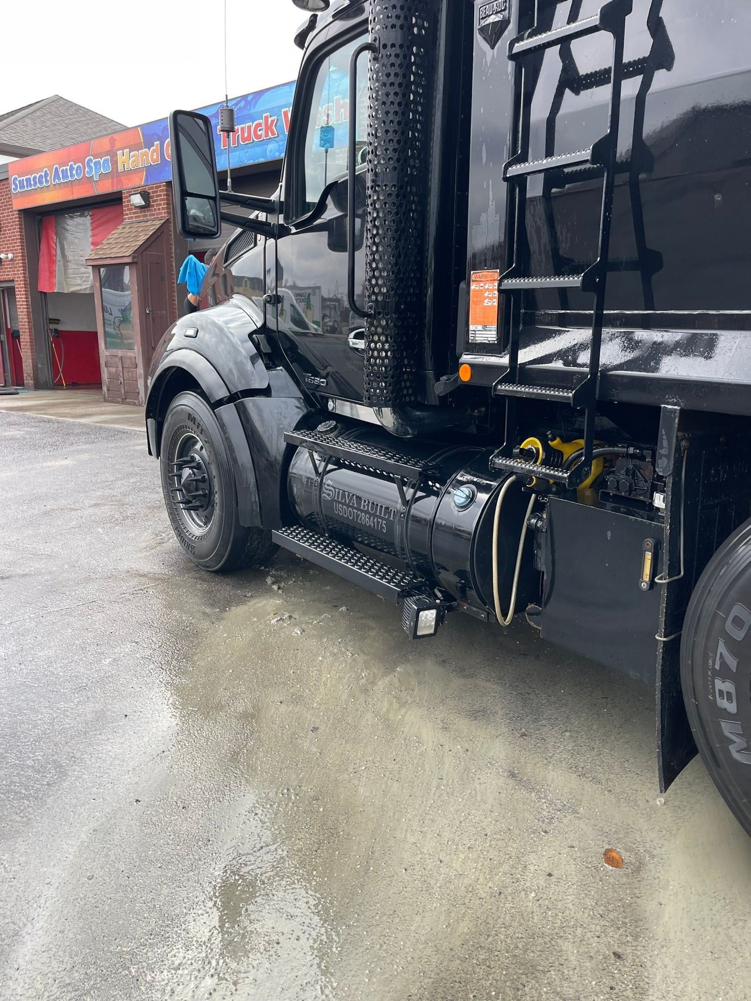 Black semi-truck parked on a wet surface, possibly after a wash, with a building in the background.