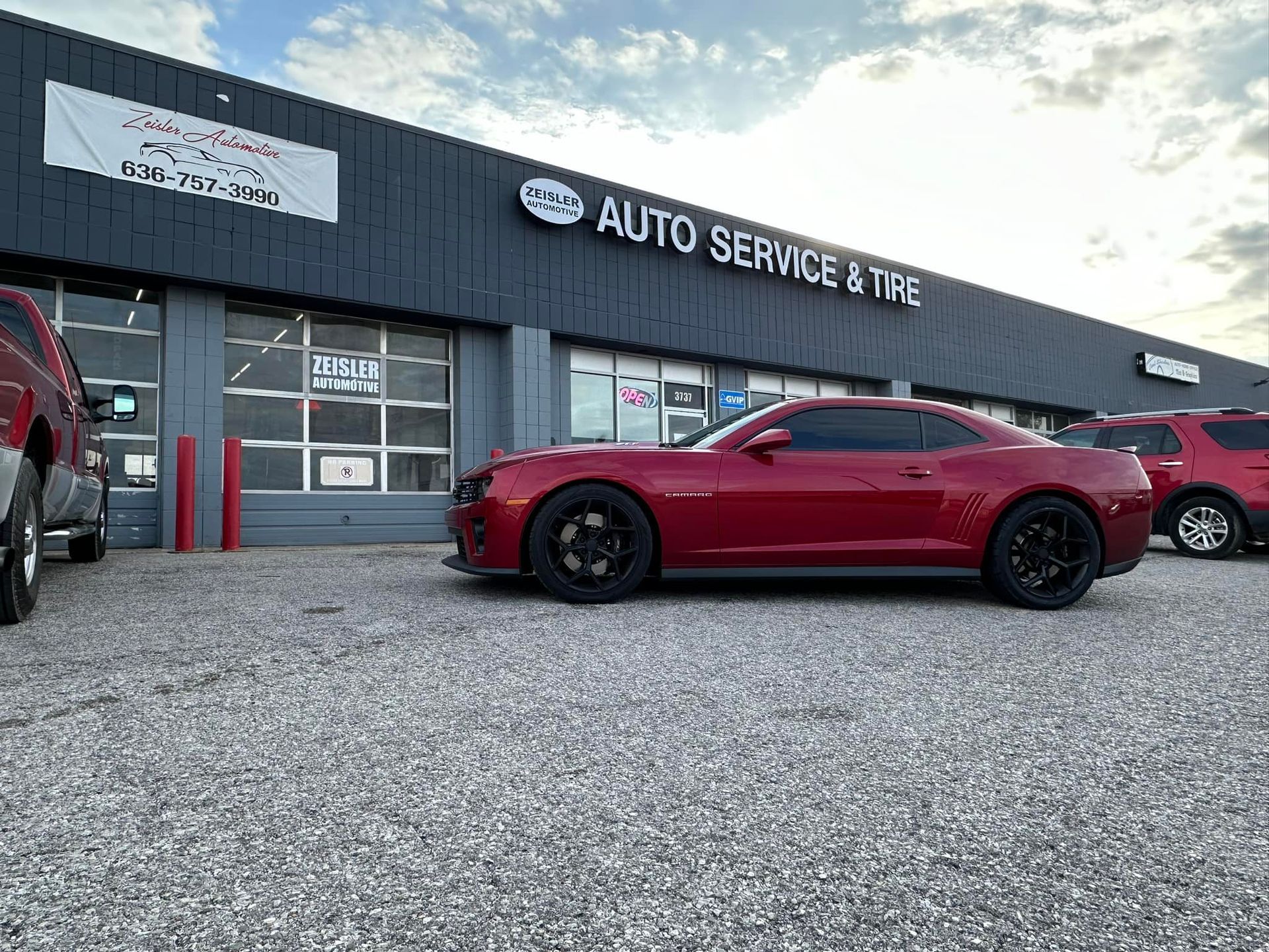 A red Chevrolet Camaro parked in front of an auto service and tire shop on a gravel lot under a cloudy sky.