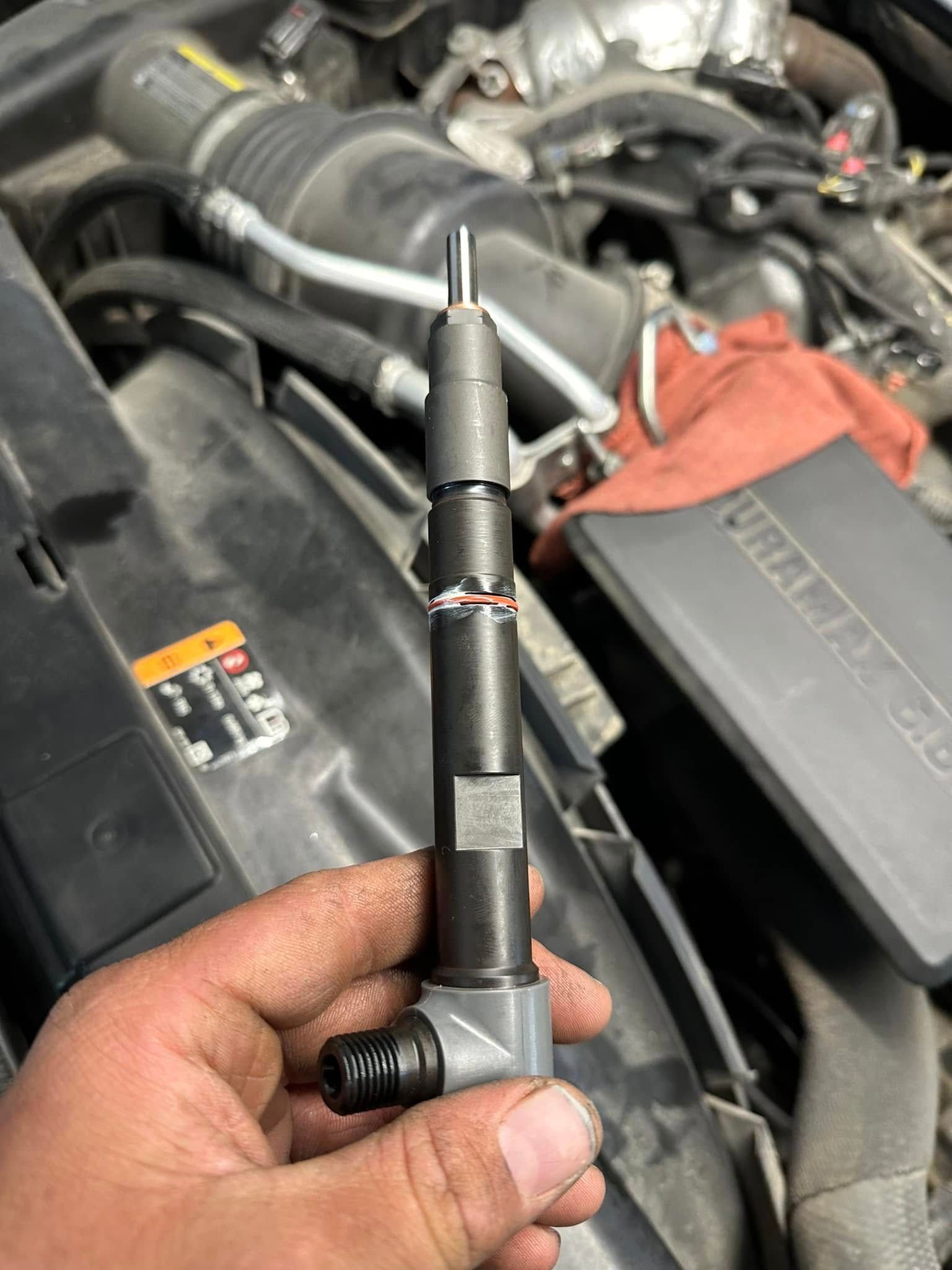 A hand holds a detached fuel injector against the background of an open vehicle engine compartment.
