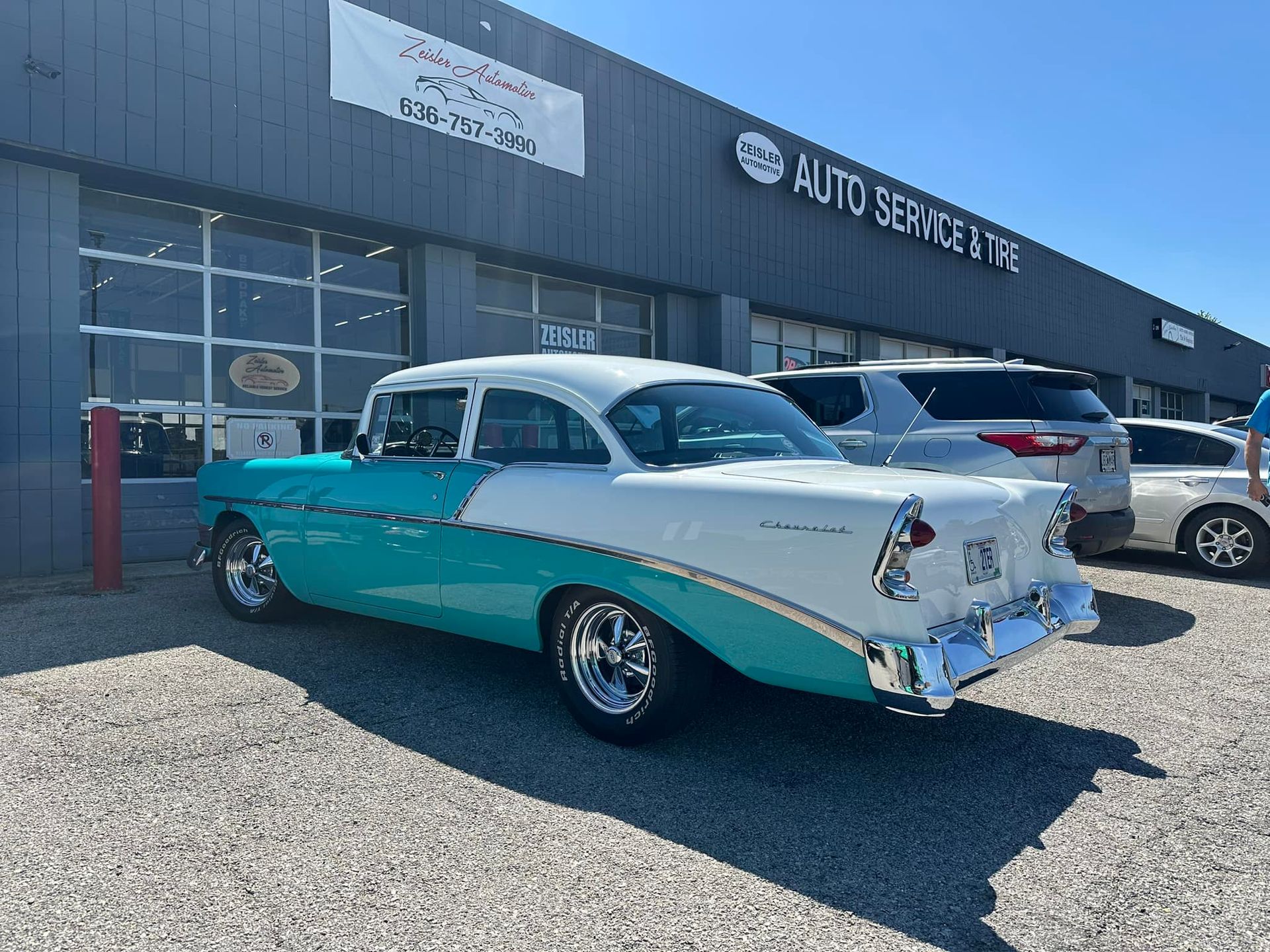 A teal and white 1956 Chevrolet Bel Air parked on a gravel lot in front of an auto service building.