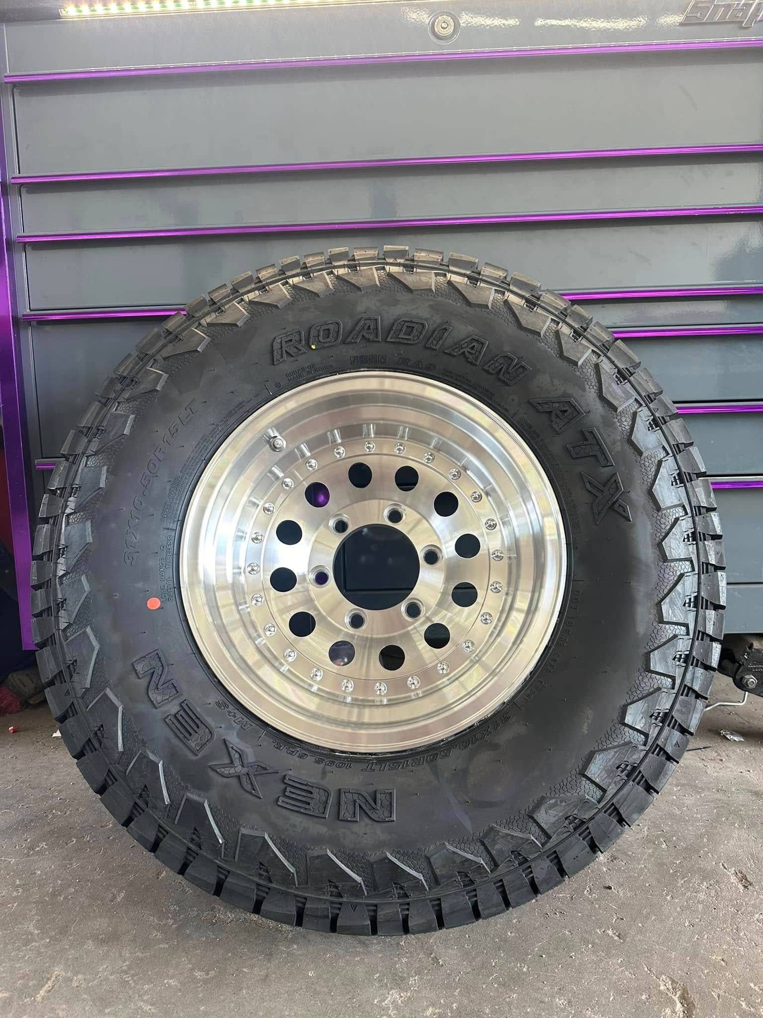 A large, black all-terrain tire mounted on a polished silver metal wheel, resting in front of a purple-accented cabinet.