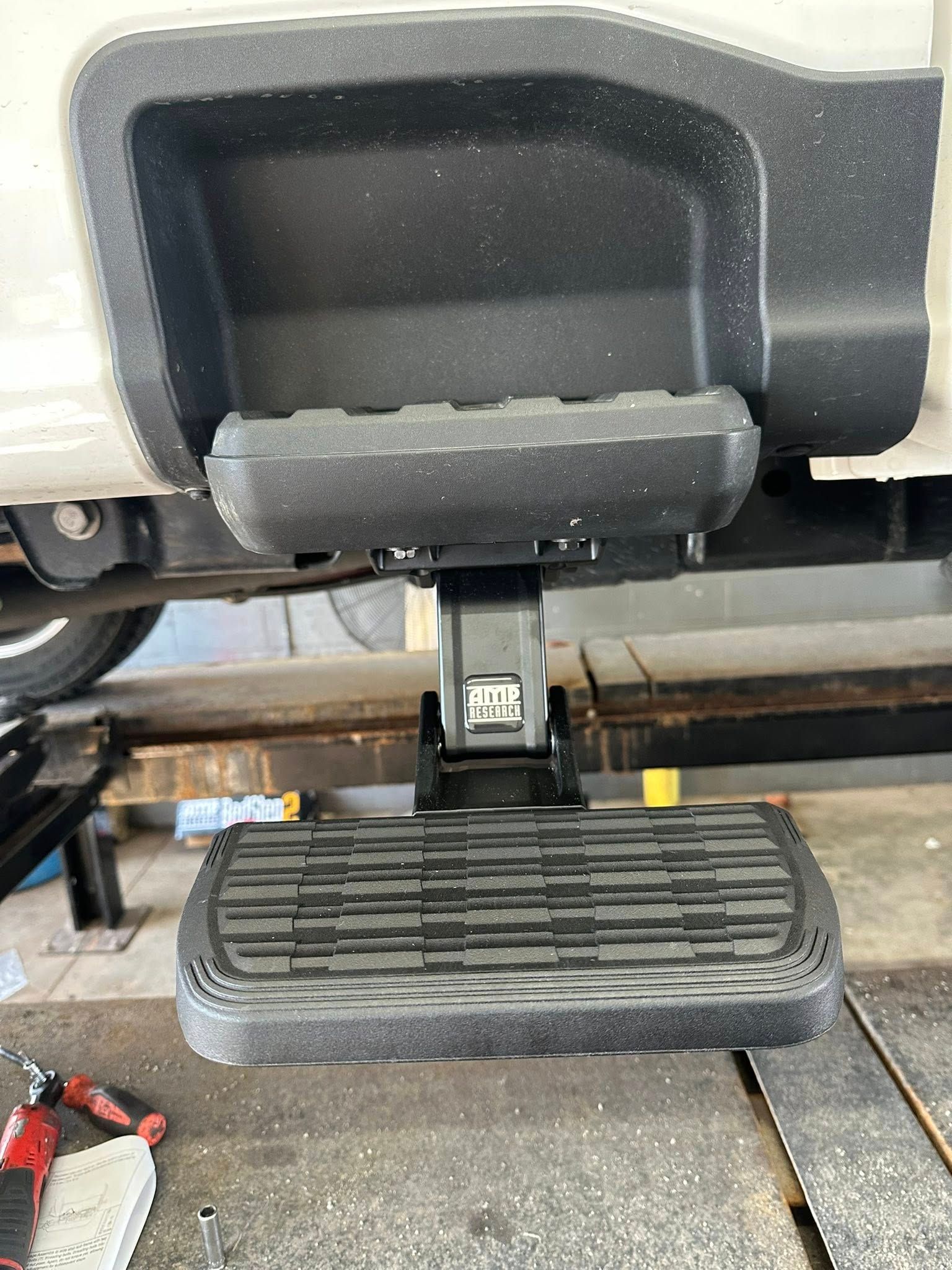 A black, retractable truck bed step mounted below a vehicle bumper, extended for access.