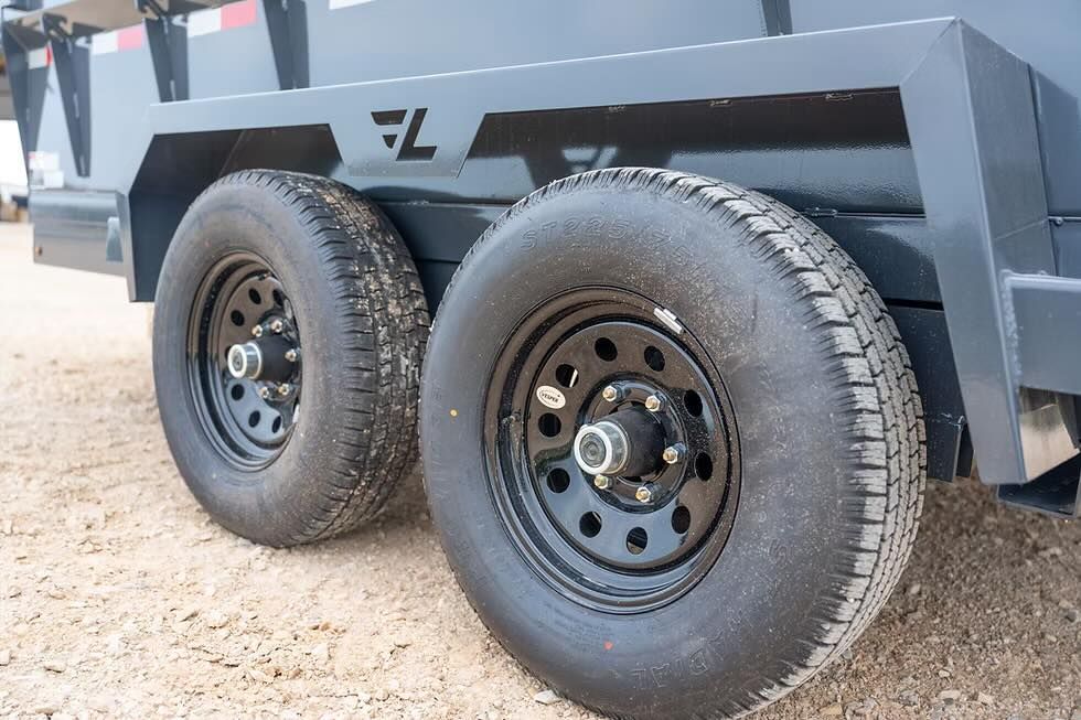 Close-up of two black trailer tires on a dark gray metal dump trailer parked on a gravel surface.