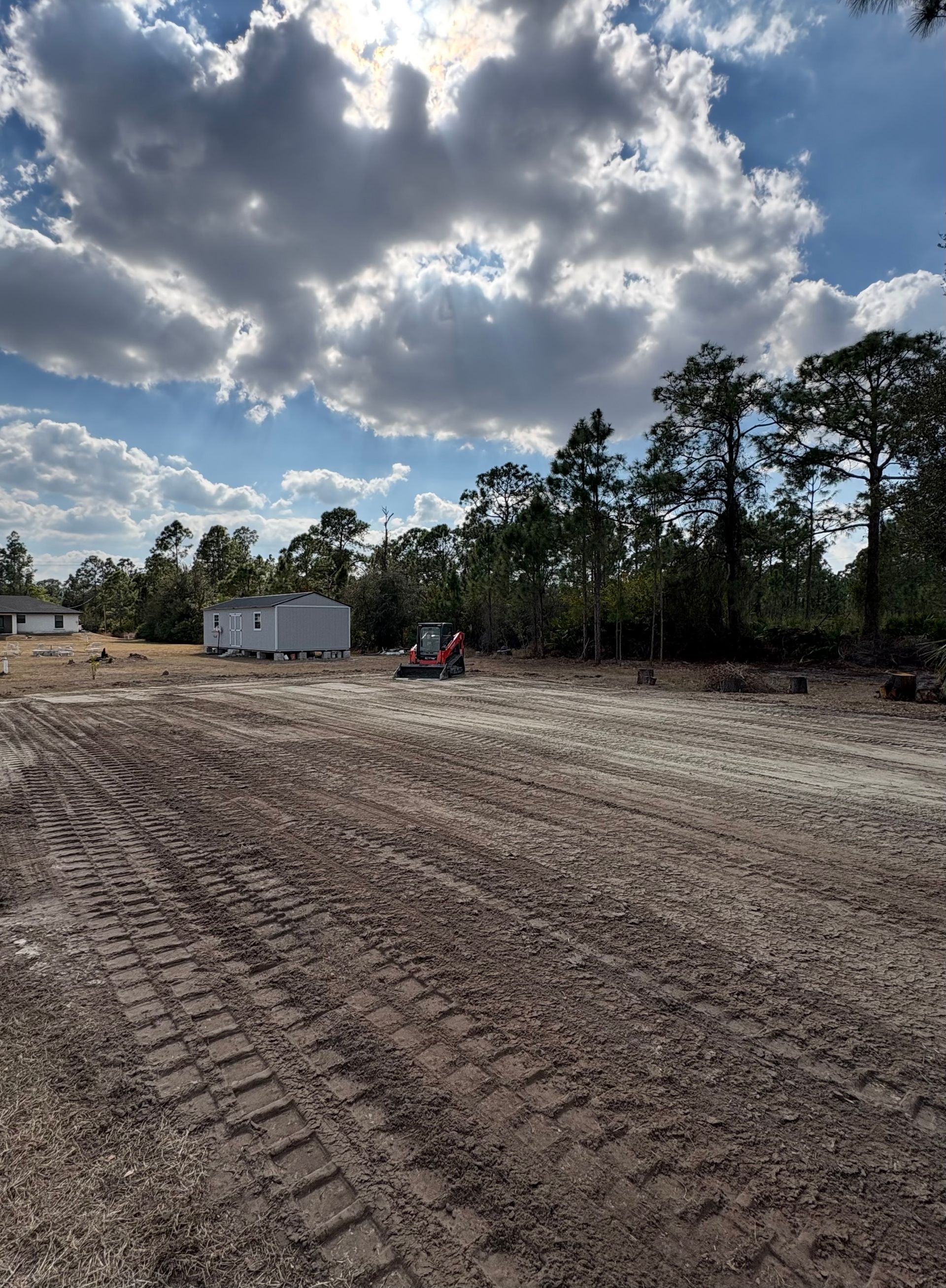 Construction site with a small building, red tractor, and dirt ground under a cloudy sky. Trees in the background.